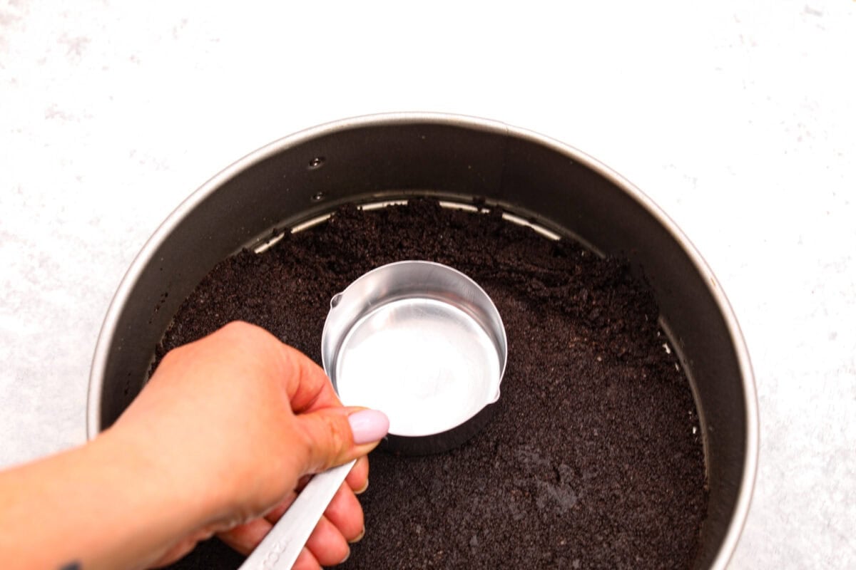 Overhead shot of someone pressing Oreo and butter mixture into a springform pan using a measuring cup. 