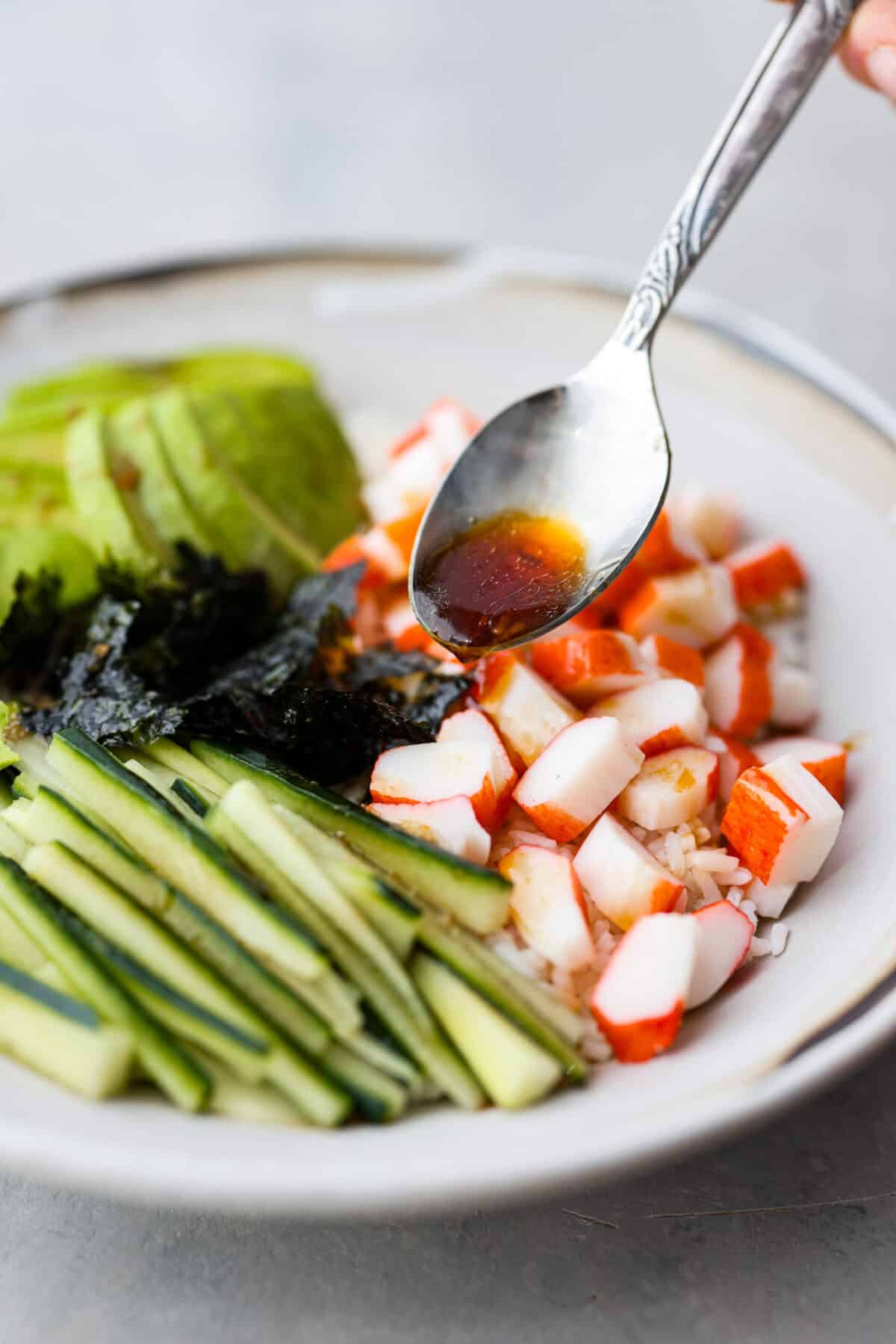 Angle shot of a plate with rice, chunks of crab, seaweed, avocado, and cucumber with poke sauce being drizzled over the top. 