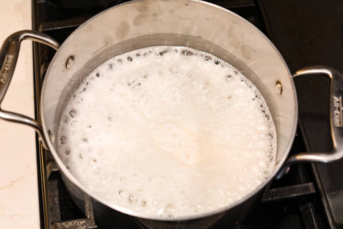 Treat yourself to this luxurious and refreshing Swedish Cream, a classic dessert that's easy to make. With a few simple ingredients and some chill time, you'll have a delightful treat topped with fresh berries and mint. Overhead shot of silver put with mixture boiling and foaming on the stovetop.