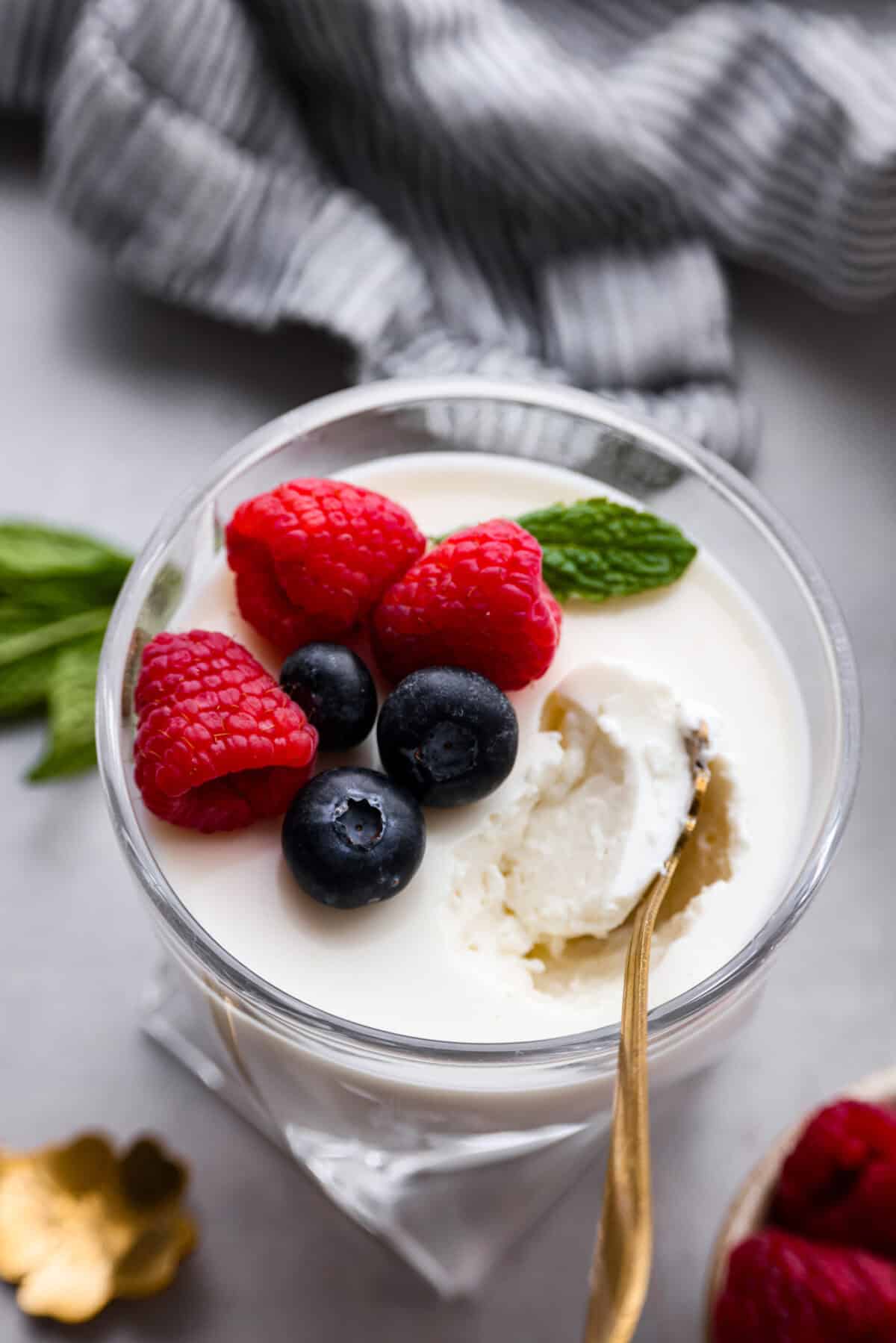 Close up shot of the top of an individual cup of Swedish cream with berries and mint on top, with a gold spoon scooping a bite out of it.  