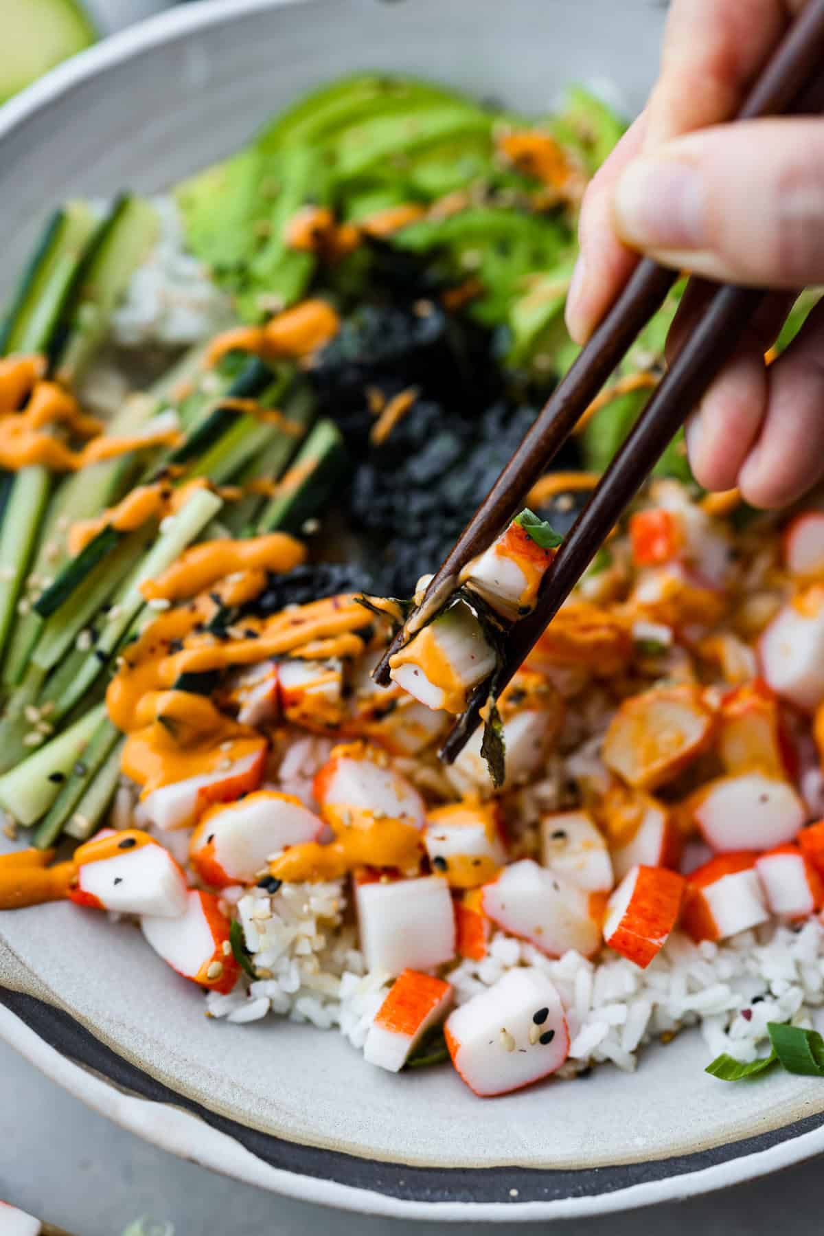 This sushi bowl is a deconstructed California sushi roll loaded with irresistible flavor and all of the best sushi ingredients. It’s layered with sushi rice, crab, cucumber, avocado, a tasty sauce, and more. It’s easy to make and ready to serve in just 20 minutes! Close up shot of someone taking a bite of the sushi bowl with chopsticks.