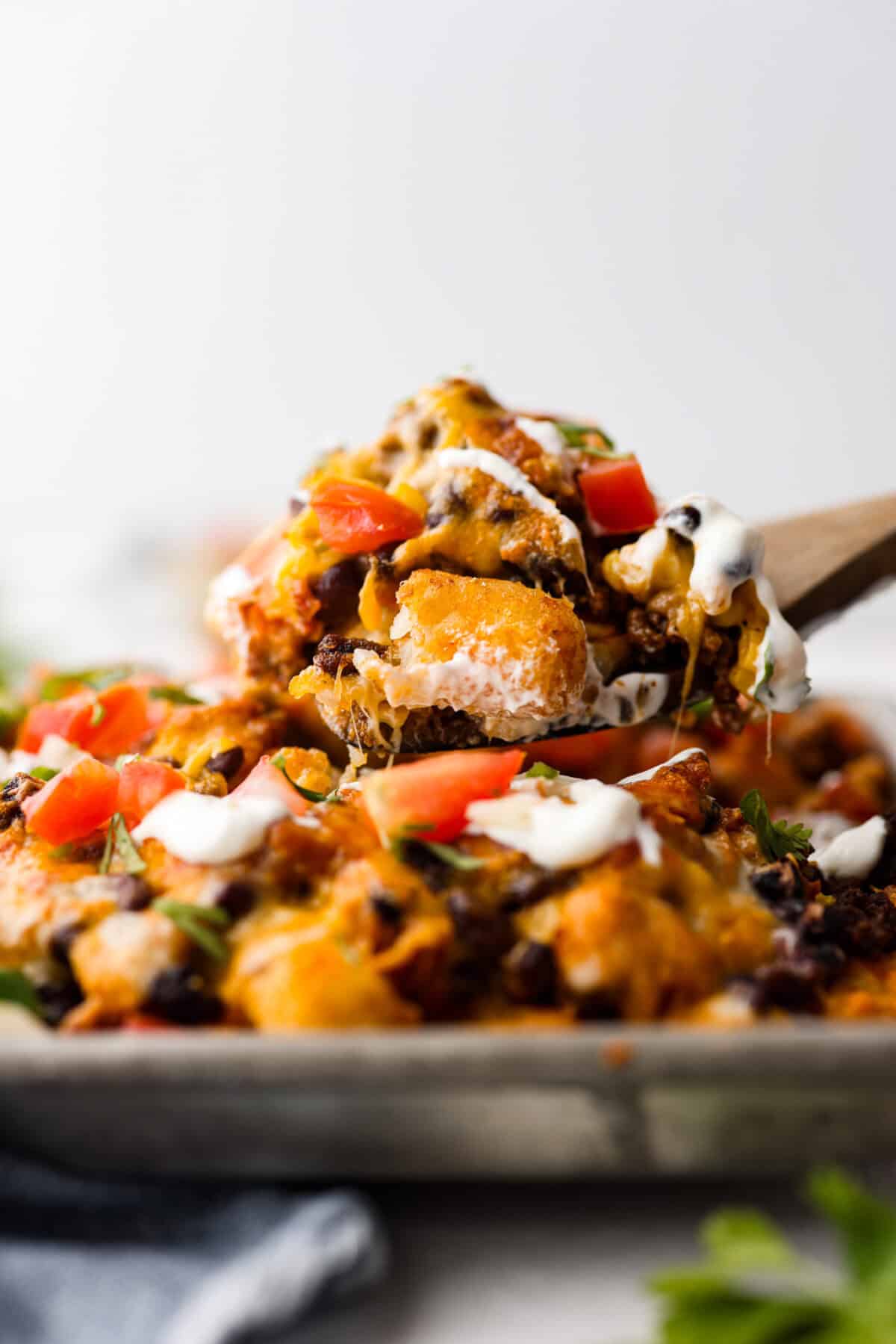 Close up side shot of a spoon full of tater tot nachos being scooped out of the pan. 