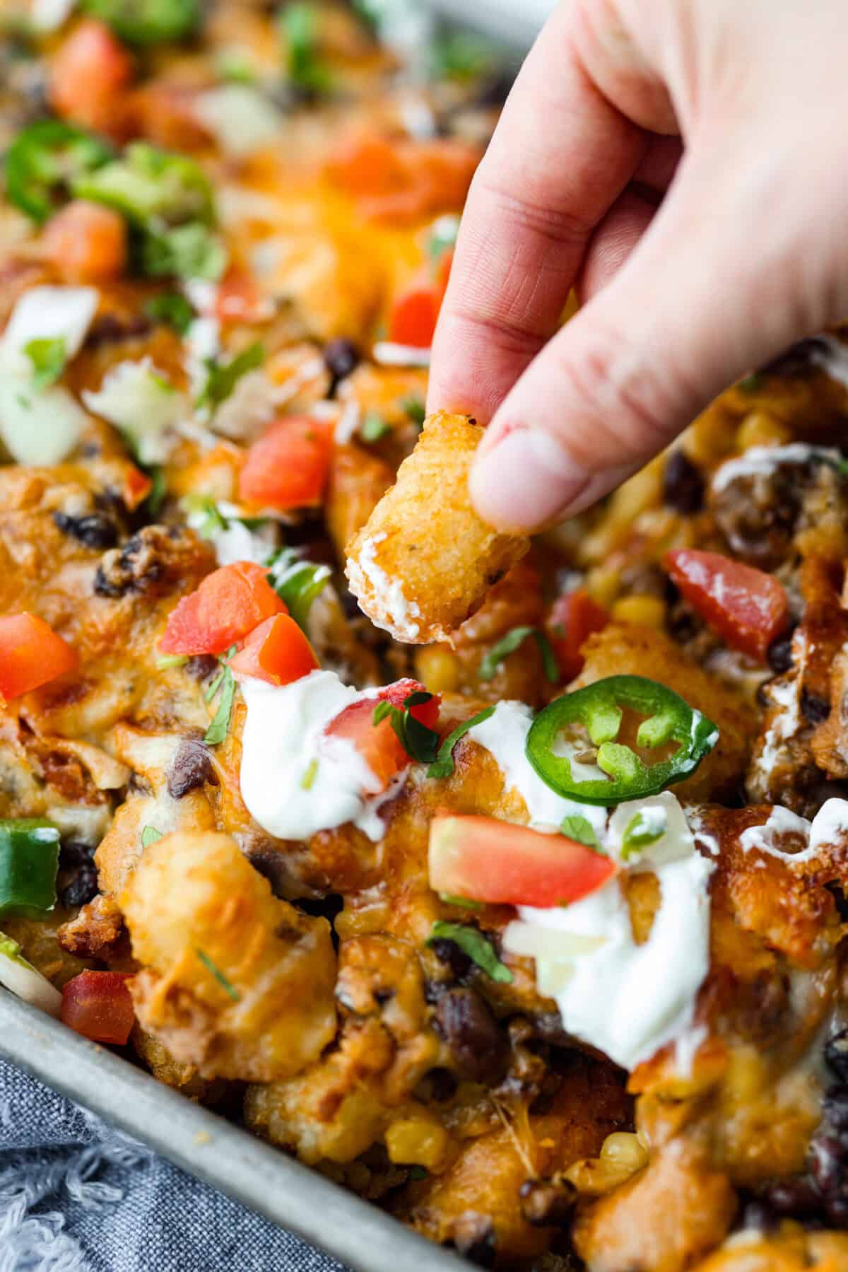 Angle shot of someone dipping a tater tot into the sour cream on that is drizzled on top of the pan of totchos. 