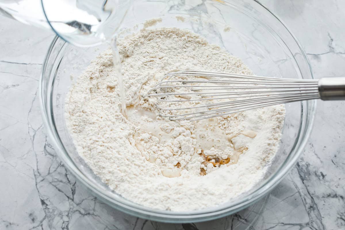 Whip up a batch of these delightful zeppole, perfect for a sweet treat anytime! With a light, fluffy texture and a generous coating of powdered or cinnamon sugar, these Italian donuts are sure to please. Overhead shot of glass bowl with mixed dry ingredients with wet ingredients being poured in.
