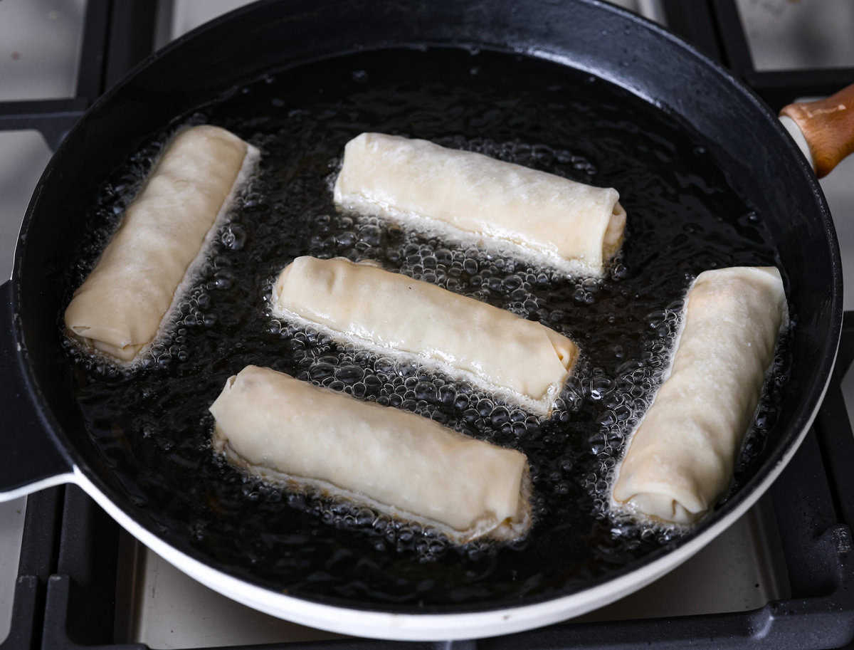 Breakfast Egg Rolls are a fun and delicious way to change up breakfast! The inside is filled with savory sausage, creamy eggs, hash browns, and plenty of cheese. Fry them up for a gooey, tender, savory center and a delicately crunchy exterior. This is a fun and unique breakfast that everyone will love! Overhead shot of 5 rolled breakfast egg rolls being fried in pan.