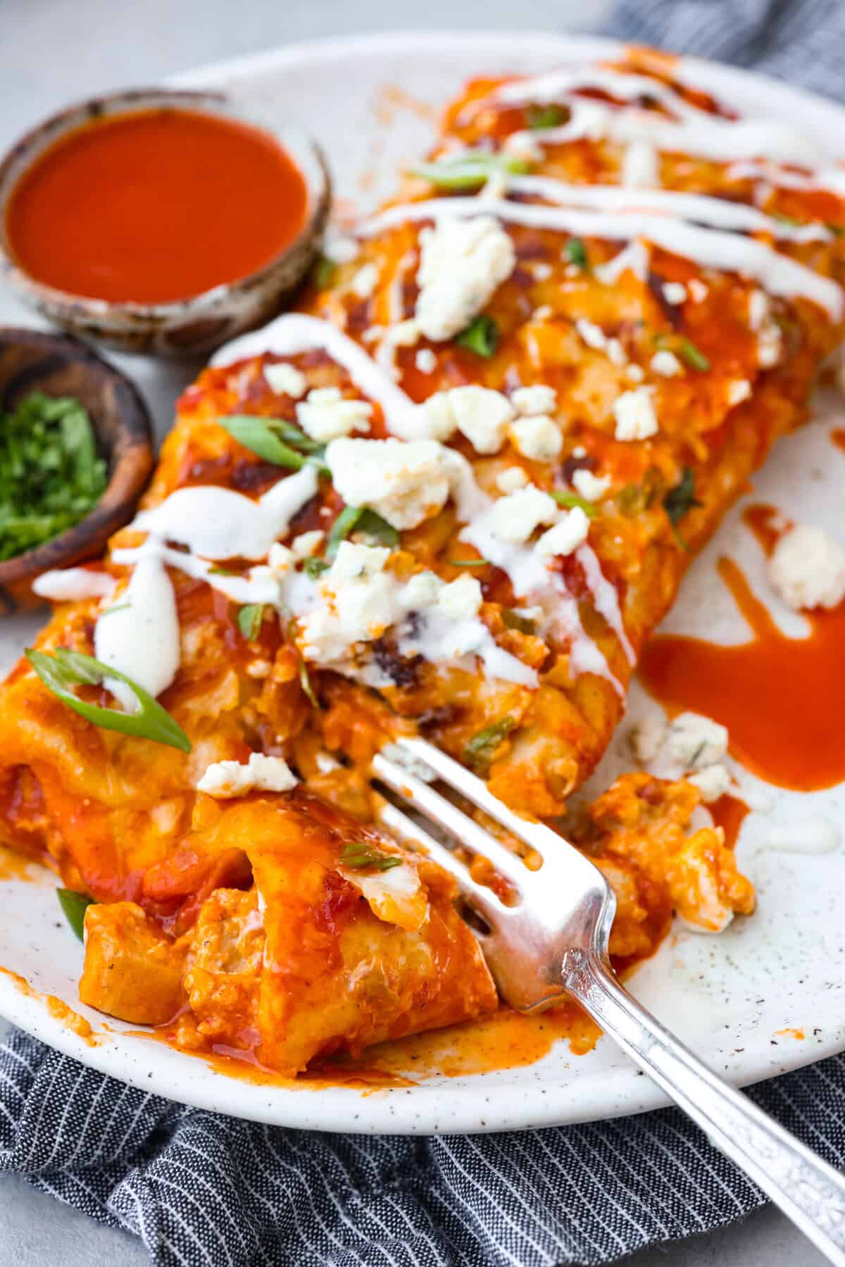 A new enchilada is in town! Get ready for the bold flavors in these buffalo chicken enchiladas! Chicken enchiladas loaded with cheese and smothered in a creamy buffalo enchilada sauce. Close view of buffalo chicken enchiladas on a plate with a fork.