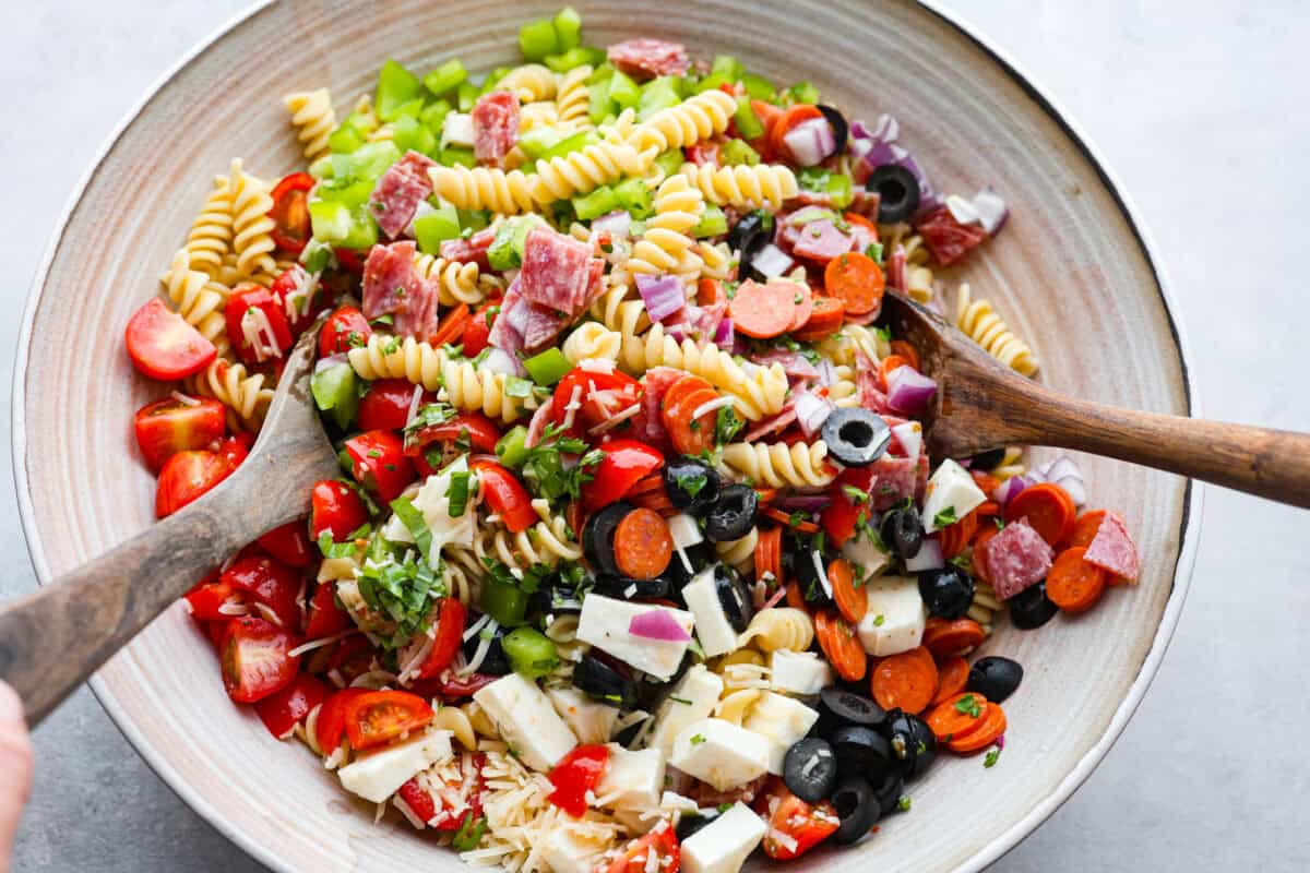 Overhead shot of large bowl with two wooden spoons stirring ingredients of pasta salad together. 