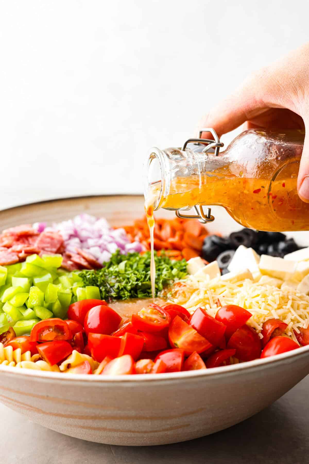 Side view of large bowl with pasta salad ingredients in piles. Someone is pouring Italian dressing into the bowl. 