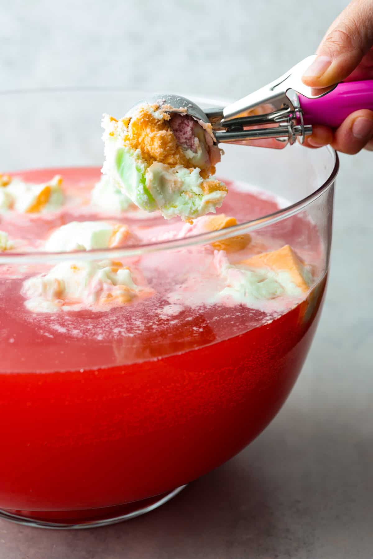 This is a picture of someone holding an ice cream scoop filled with rainbow sherbet over a large bowl filled with the drink and other dollops of rainbow sherbet. 