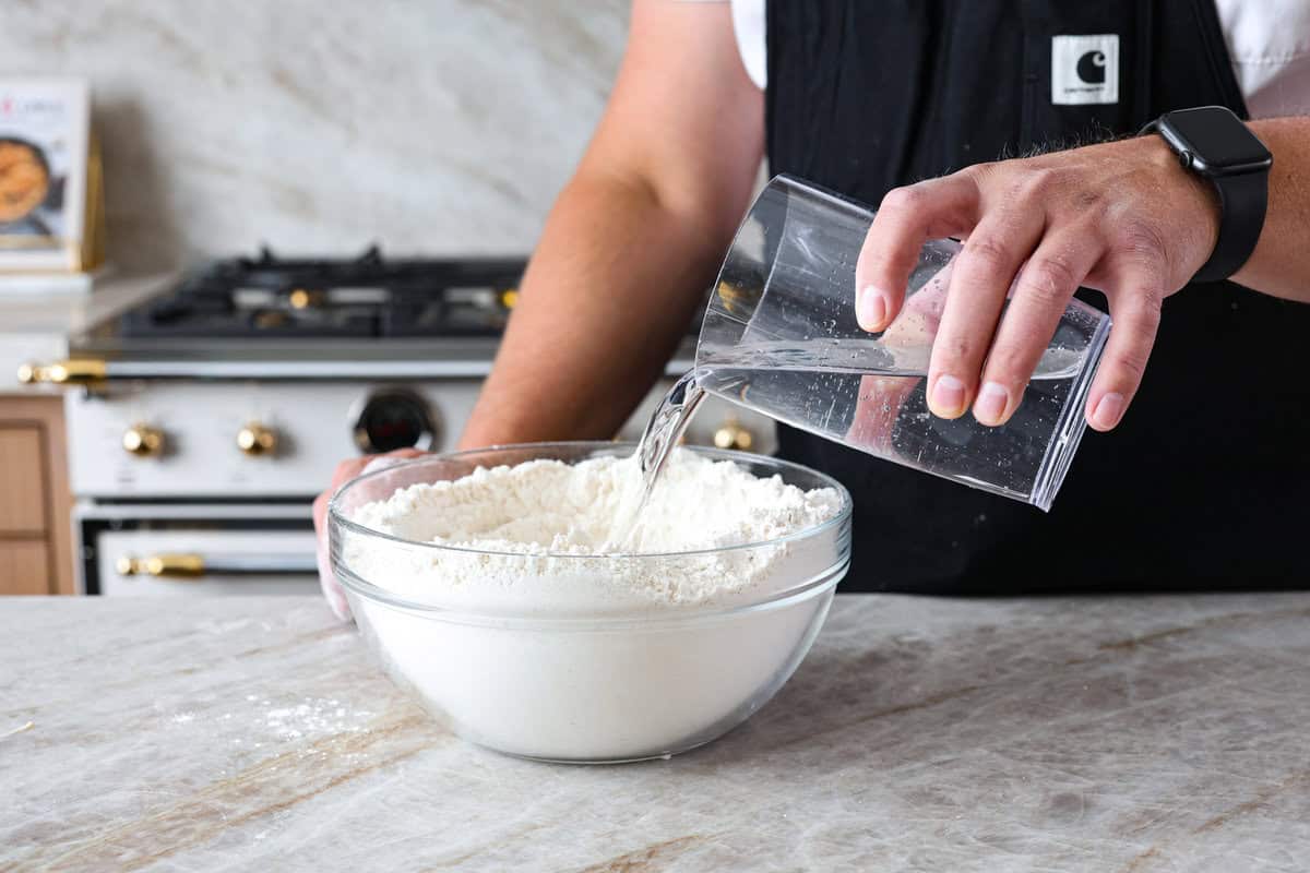 This is the perfect beginner's guide for making wood-fired pizza dough! Pizza expert, Chef Brandon Price, taught me how to make authentic homemade pizza, and now you get to learn too! Third photo of the water pouring into the bowl to make the crust.