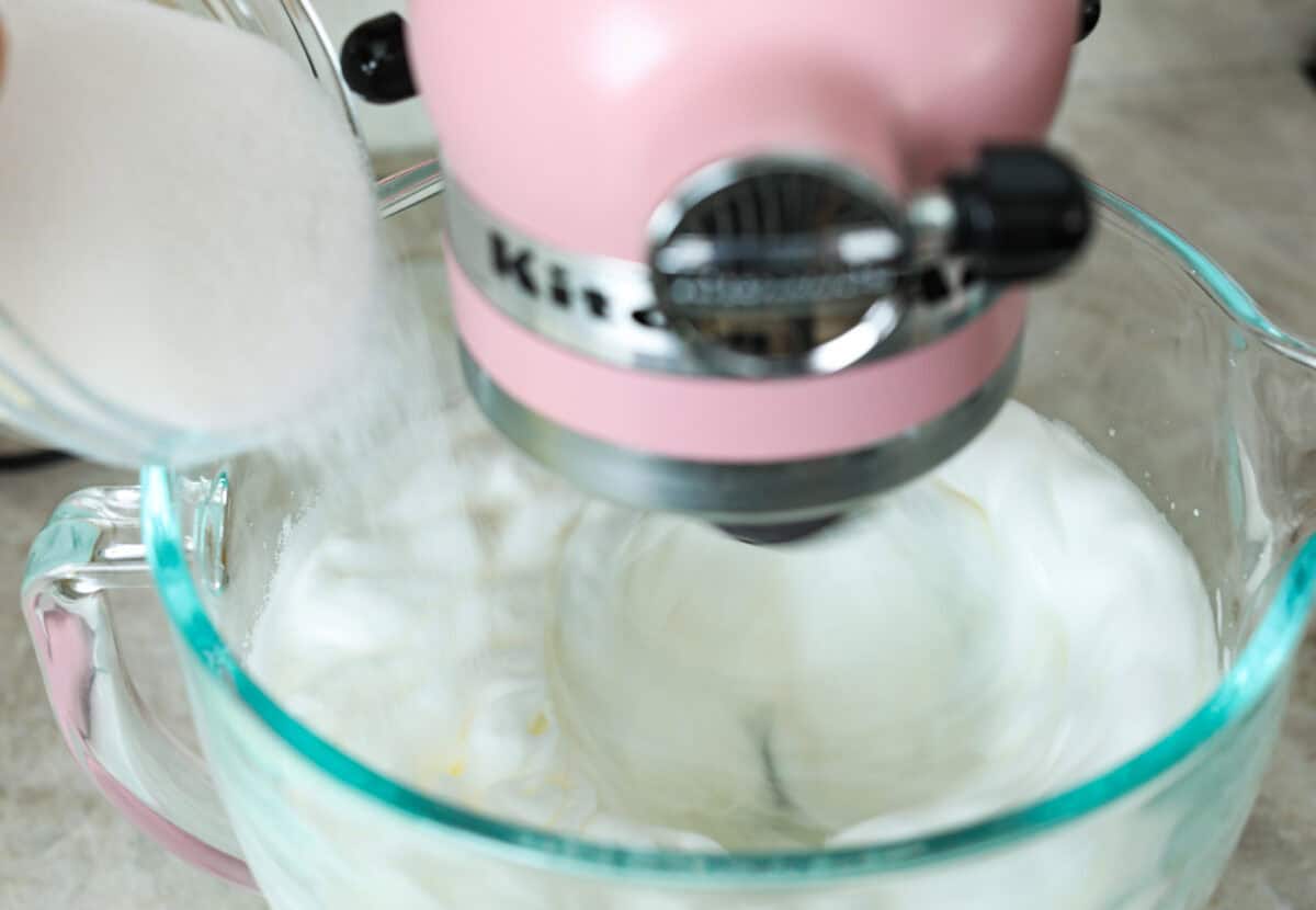 Angle shot of sugar being poured into  stand mixer bowl full of whipped egg whites. 