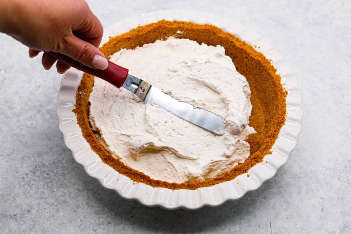 Overhead shot of someone smoothing pie filling with metal spatula into graham cracker crust.