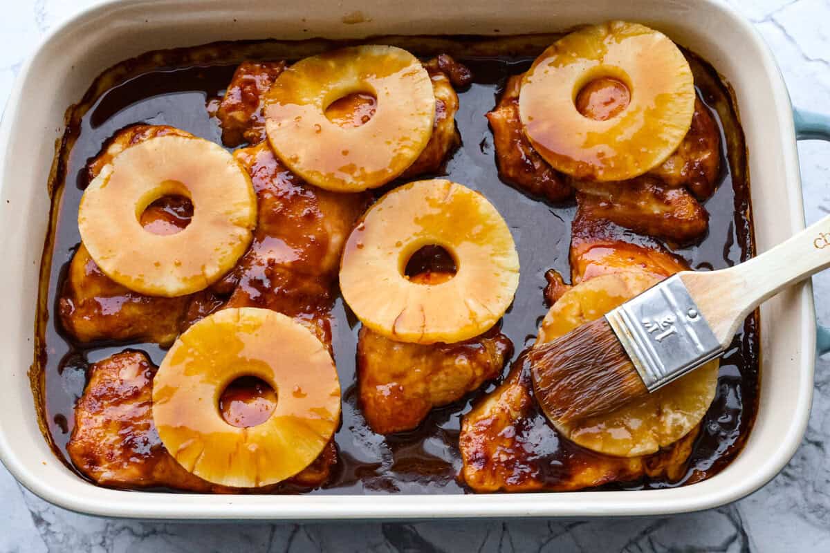 Baked Huli Huli chicken is a tried and true recipe now made in the oven! Tender chicken baked in a sweet and savory sauce. This easy one-pan dinner is full of Hawaiian-inspired flavors! Fourth photo of the pineapple placed on top of the chicken and the sauce basting on top.