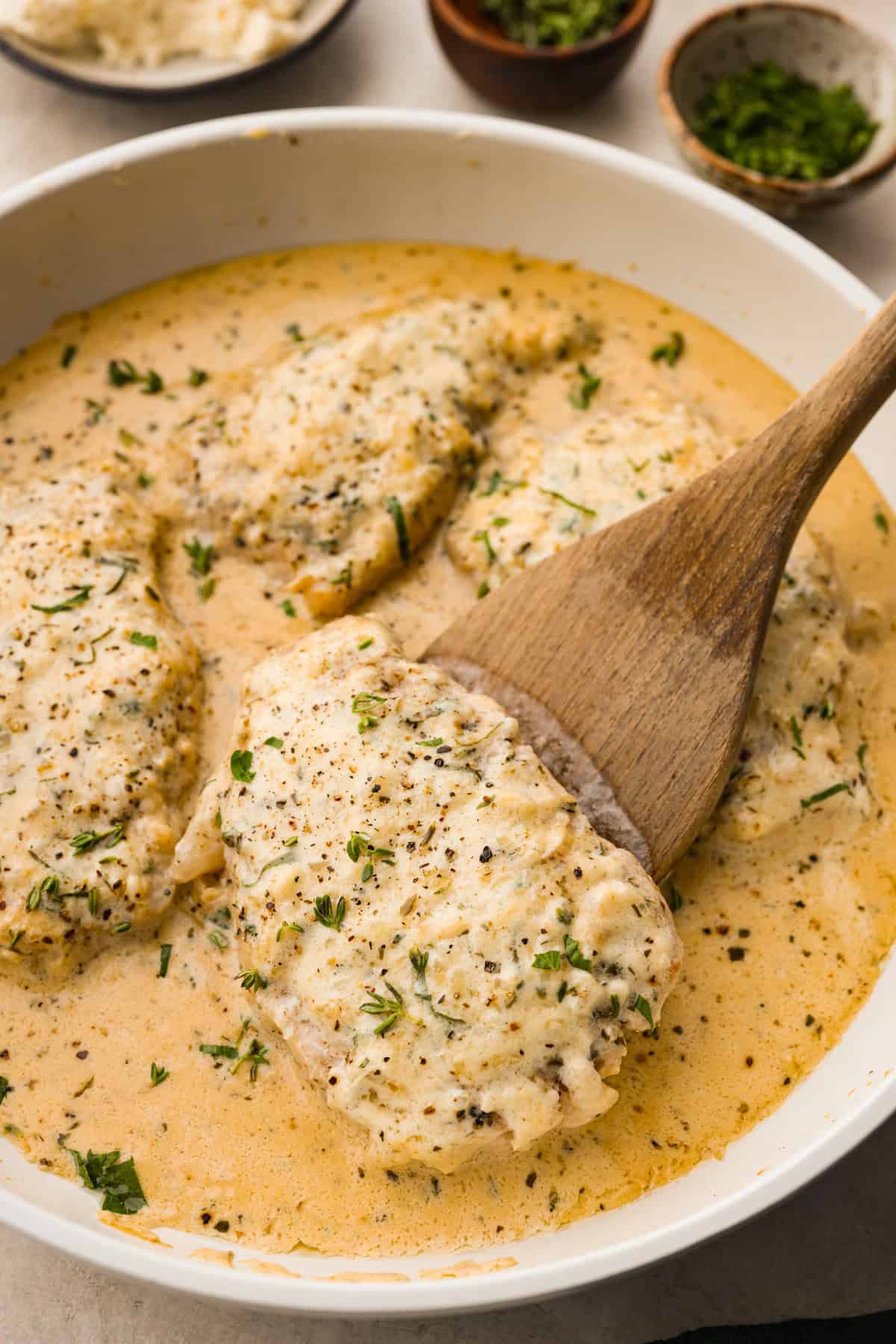 Boursin Pork Chops are savory, tender, and covered in a delicious cheesy cream sauce. This one-pan recipe is the perfect easy main that can be on your dinner table in just 30 minutes! Angle shot of wooden spoon dishing out a Boursin pork chop out of pan.