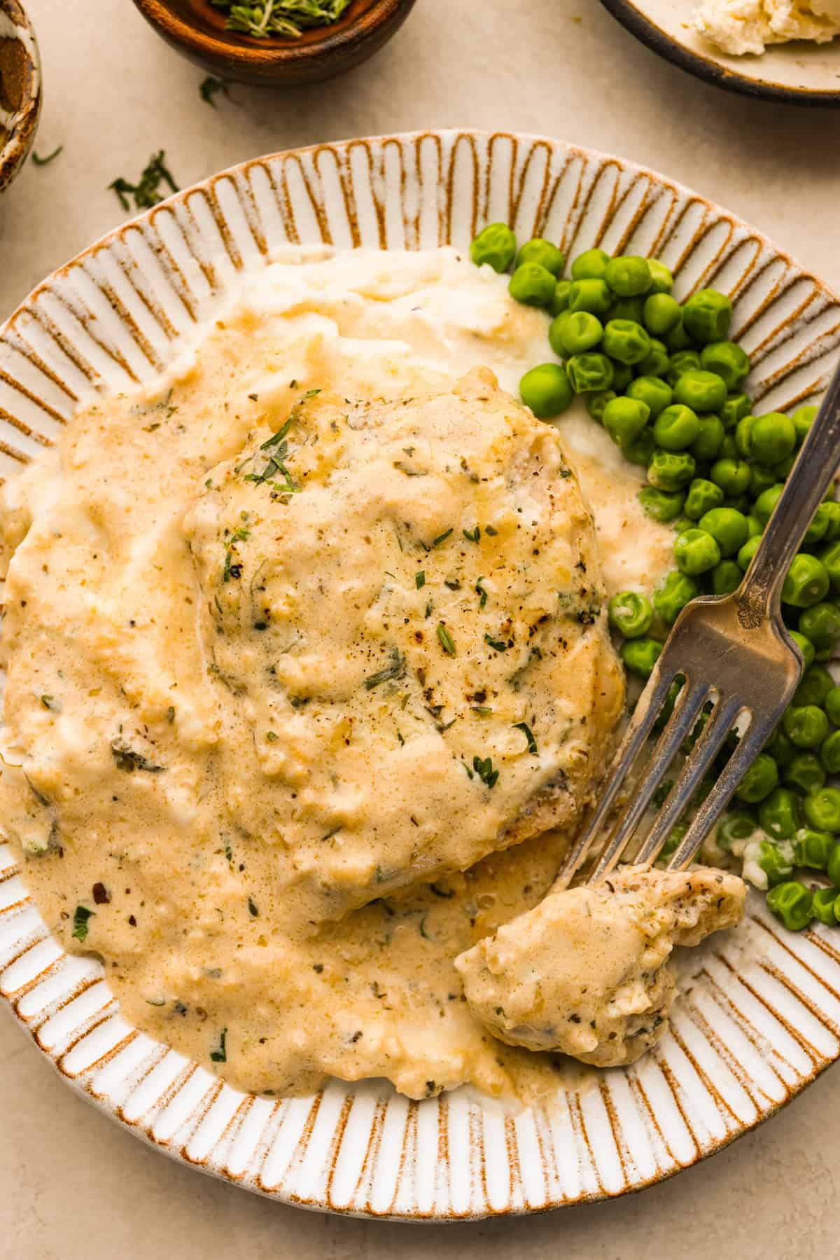 Boursin Pork Chops are savory, tender, and covered in a delicious cheesy cream sauce. This one-pan recipe is the perfect easy main that can be on your dinner table in just 30 minutes! Overhead shot of plated Boursin pork chop with peas on the side and a bite skewered on a fork.