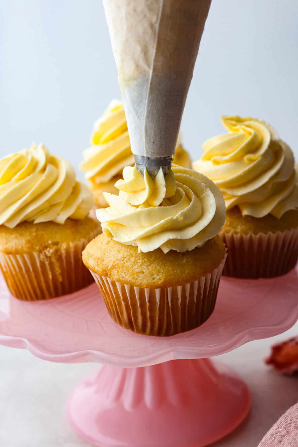 Side shot of french buttercream being piped onto a cupcake that is on a pink cake stand with other cupcakes. 