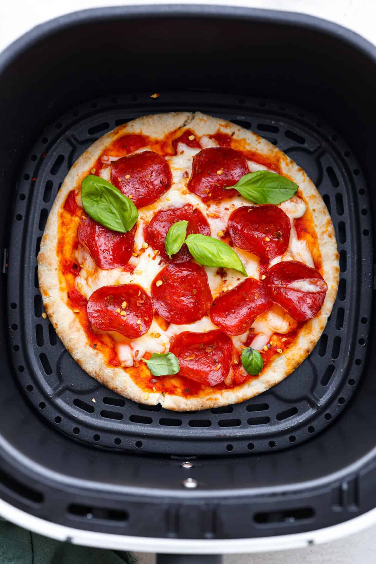 Overhead shot of cooked pita pizza in the bottom of air fryer basket garnished with pepper flakes and basil. 