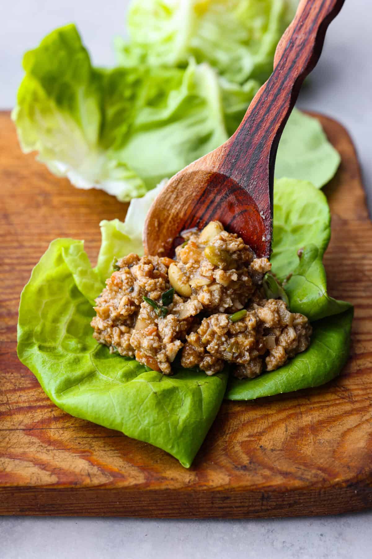 Close up shot of wooden spoon placing a scoop of lettuce wrap filling into a lettuce leaf. 