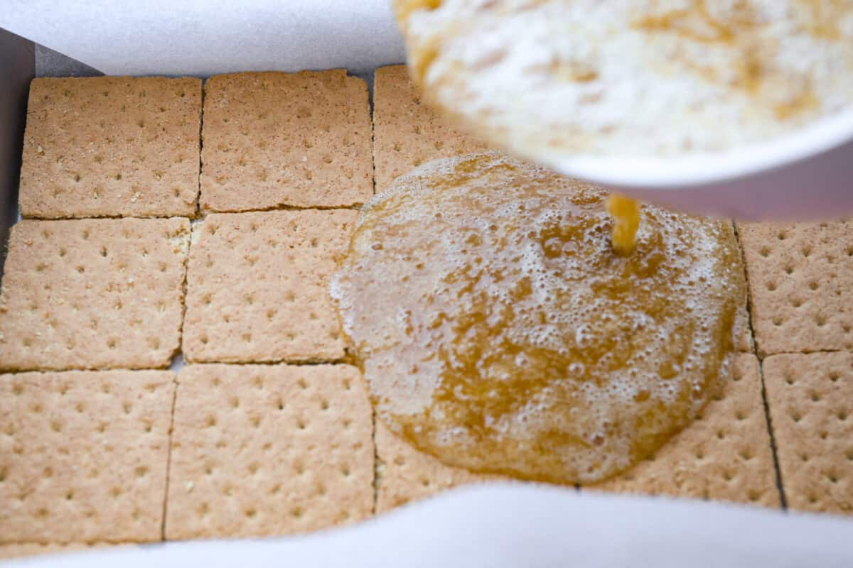 A bowl of melted butter and sugar mixture being poured over the top of the pan with the layer of graham crackers. 
