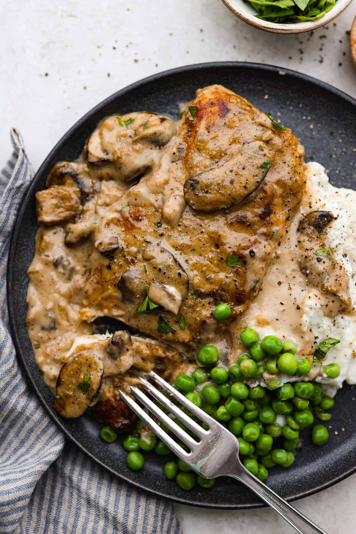 Smothered chicken is a flavorful comfort food full of rich, creamy gravy, tender chicken cutlets, and savory mushrooms. Perfect on its own or paired with mashed potatoes, rice or pasta, you're family will definitely want this on the weekly meal plan. Overhead shot of plated smothered chicken over mashed potatoes and a side of peas.