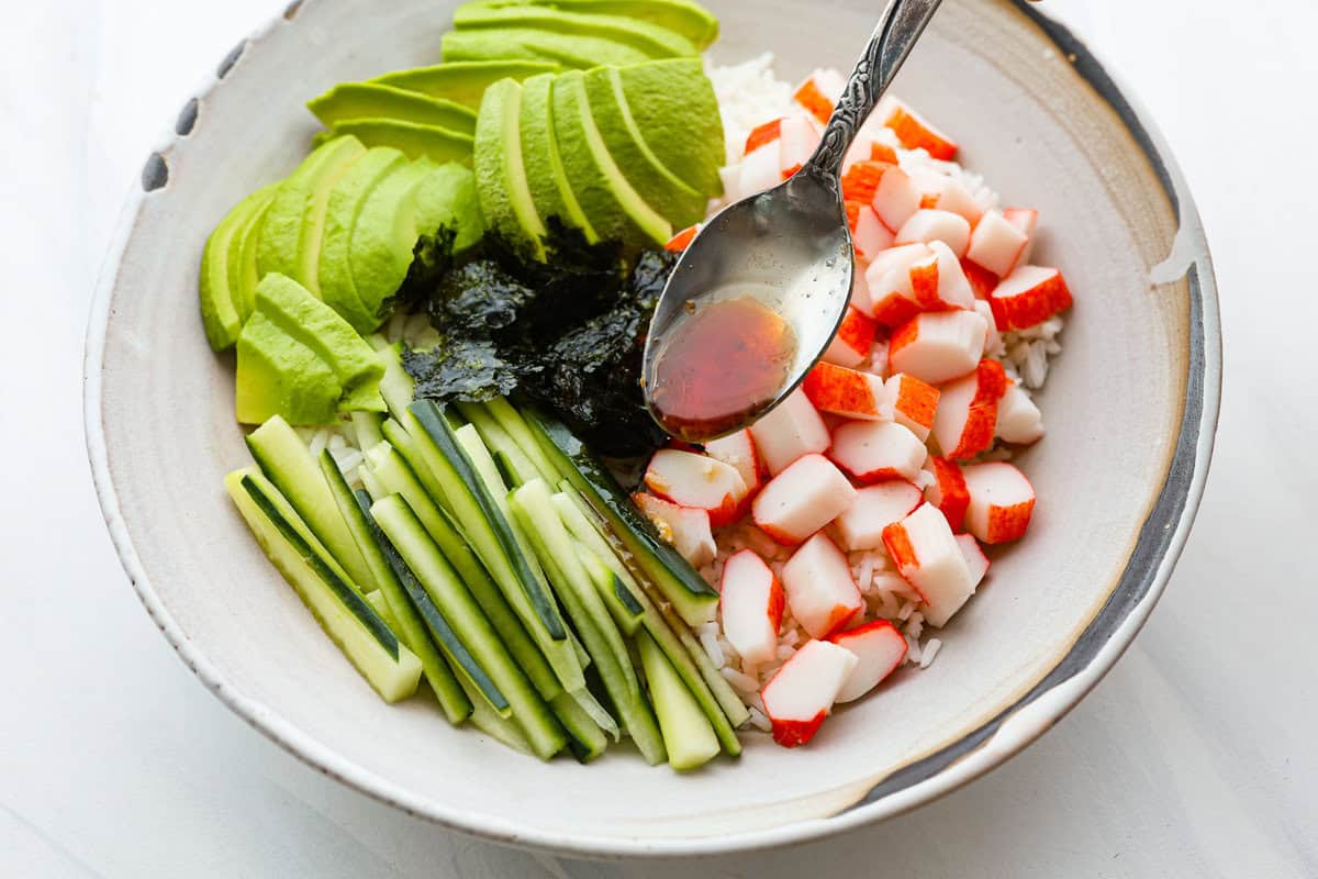This sushi bowl is a deconstructed California sushi roll loaded with irresistible flavor and all of the best sushi ingredients. It’s layered with sushi rice, crab, cucumber, avocado, a tasty sauce, and more. It’s easy to make and ready to serve in just 20 minutes! Overhead shot of sushi bowl with someone pouring sauce over it with a spoon.