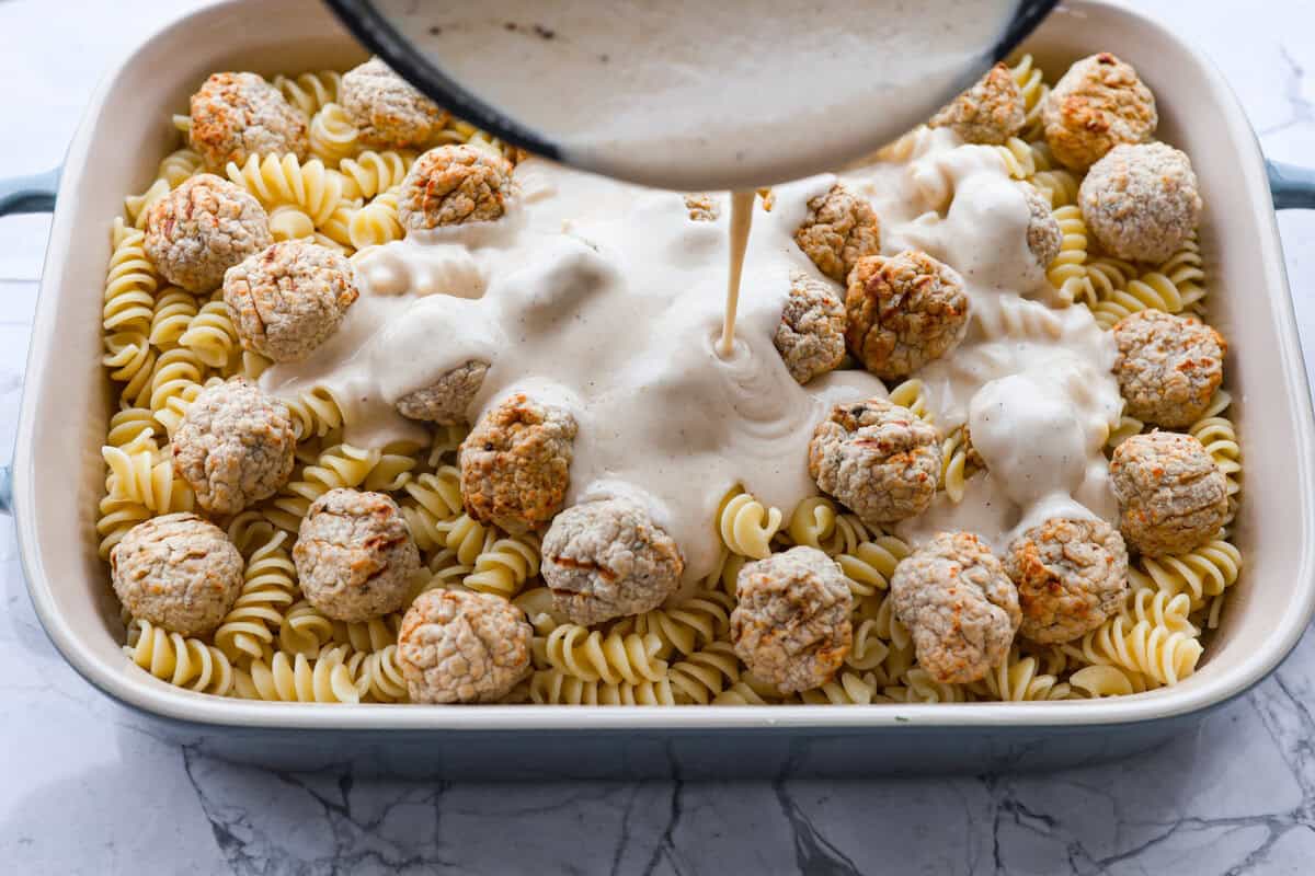 Swedish Meatball Casserole is a classic comfort meal the whole family will enjoy! Tender meatballs are layered over perfectly cooked pasta and baked in a rich, creamy gravy. This dinner recipe is easy enough to make during the week but also deliciously impressive for entertaining! Overhead shot of someone pouring sauce over pasta and meatballs in a casserole dish.