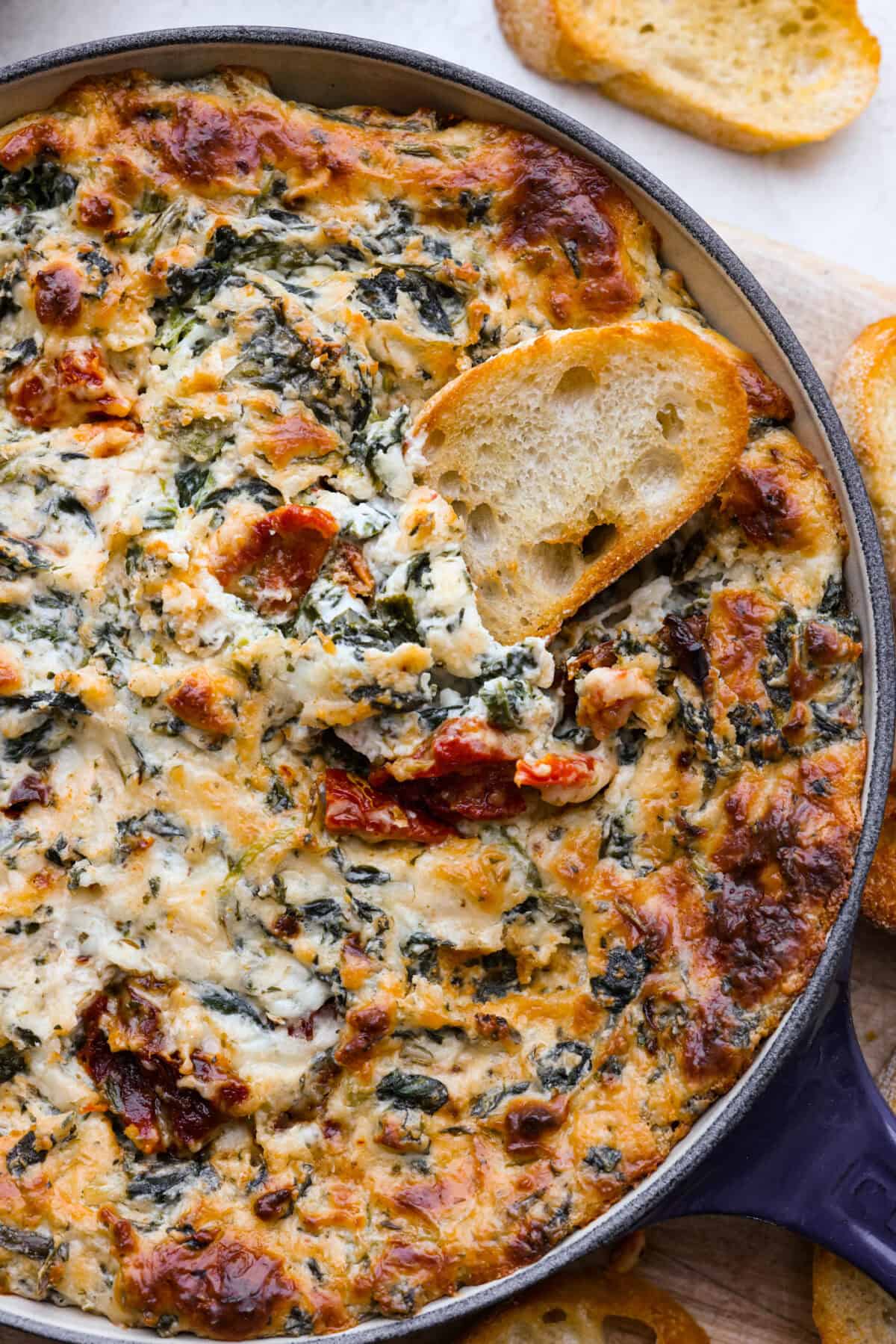 Close top view of creamy Tuscan dip in a blue skillet with a crostini dipping in.