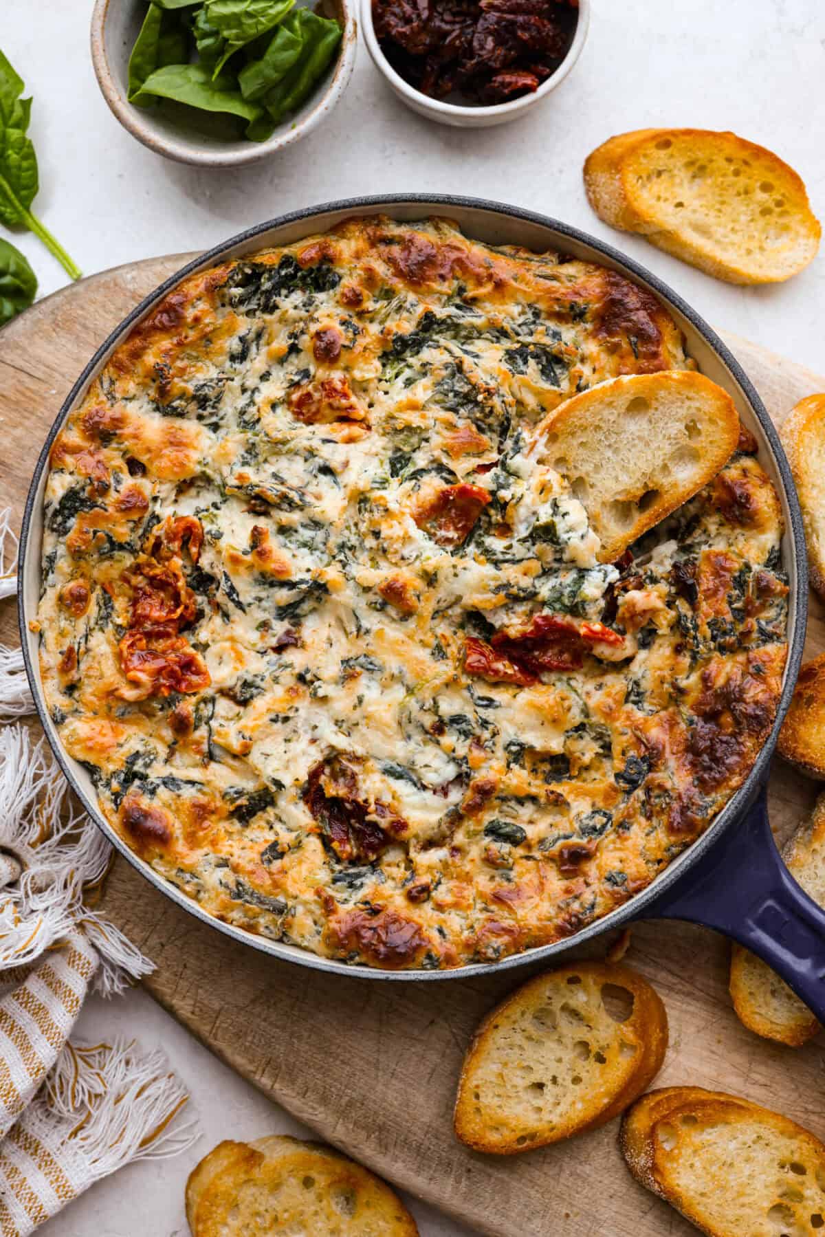 Top view of creamy Tuscan dip in a skillet with crostini scattered on a wood board.