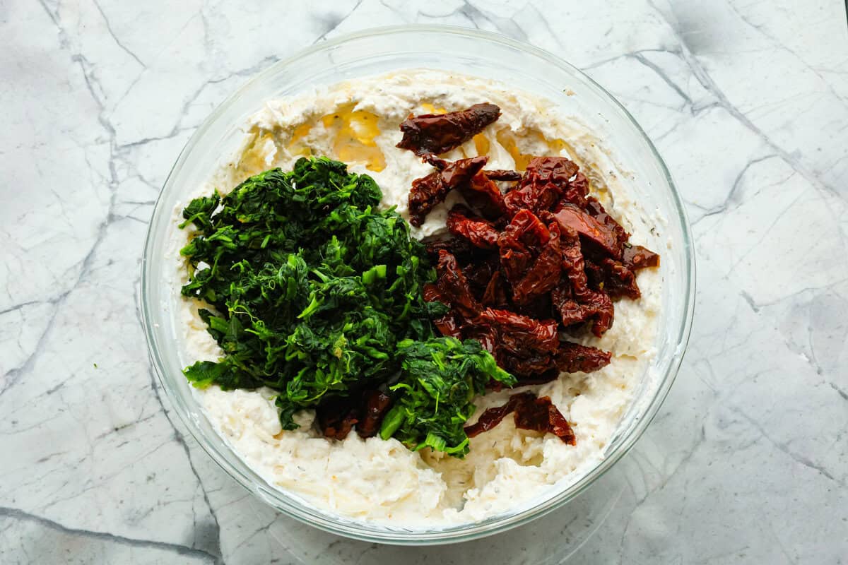 There's a new appetizer in town, and this creamy Tuscan dip won everyone's hearts! The combination of cheeses, garlic, spinach, and sun-dried tomatoes creates an addicting dip you’ll want to bring to all of your gatherings! Third photo of the spinach and sun-dried tomatoes added to the bowl.