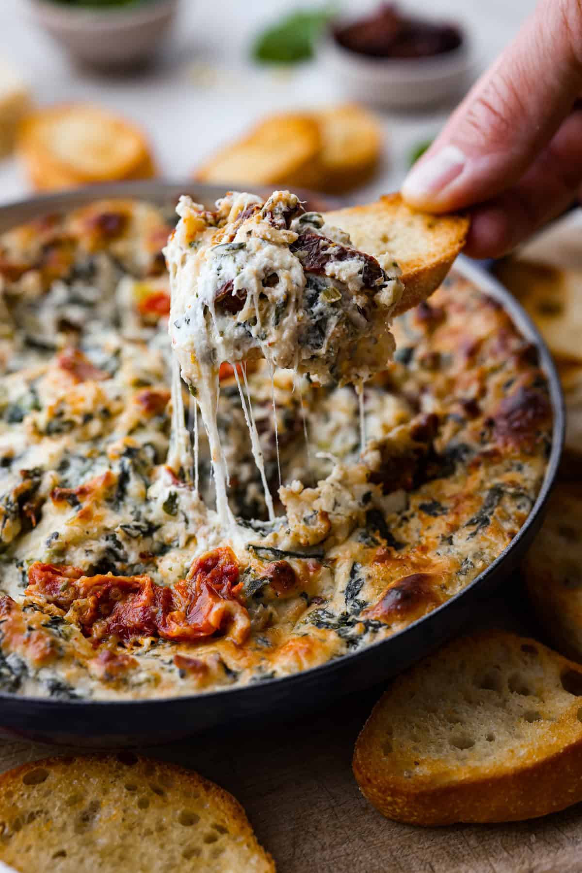 There's a new appetizer in town, and this creamy Tuscan dip won everyone's hearts! The combination of cheeses, garlic, spinach, and sun-dried tomatoes creates an addicting dip you’ll want to bring to all of your gatherings! Close view of crostini dipping into a the dip with a cheese pull.
