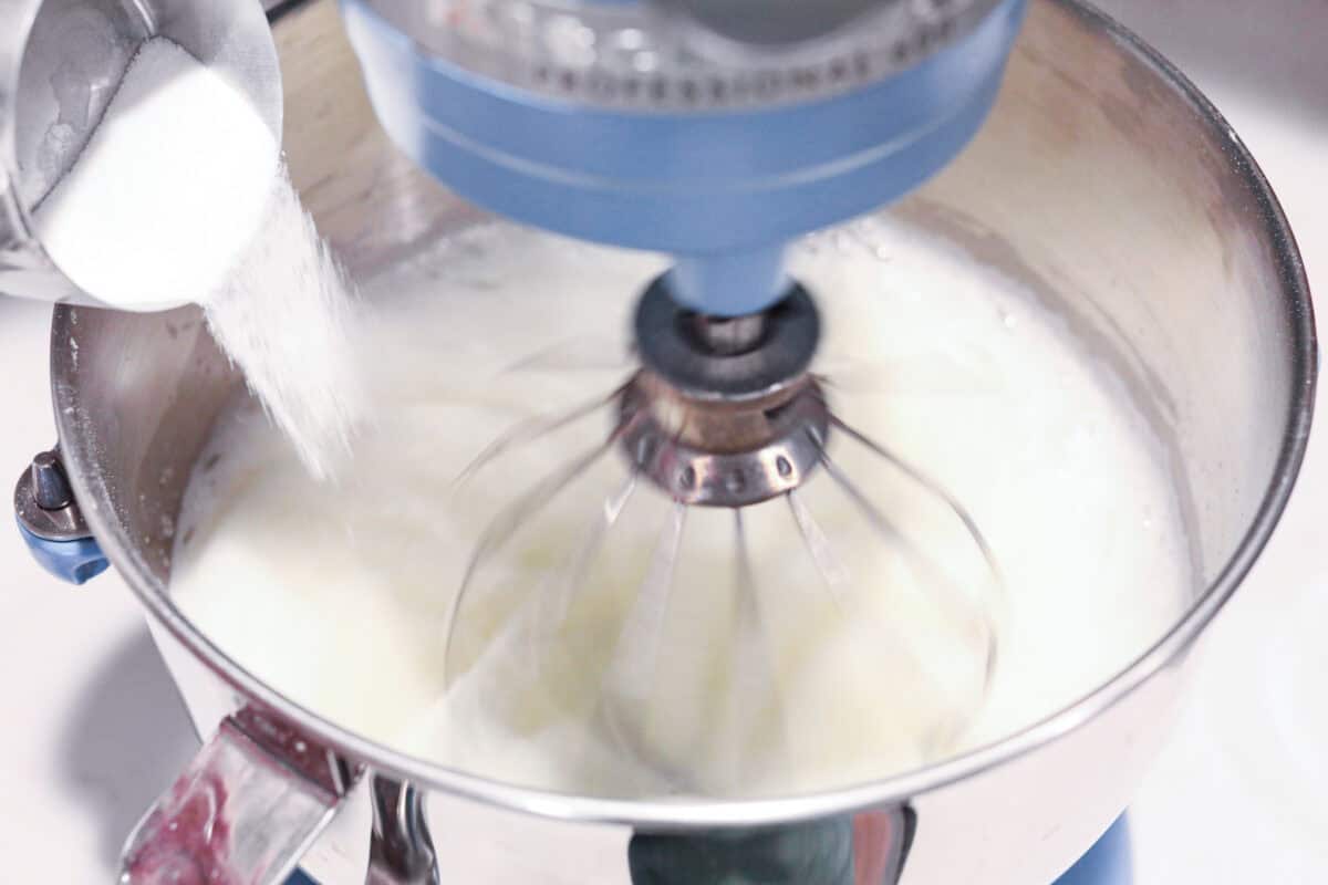 Overhead shot of sifted sugar being poured into a stand mixer whisking egg whites. 