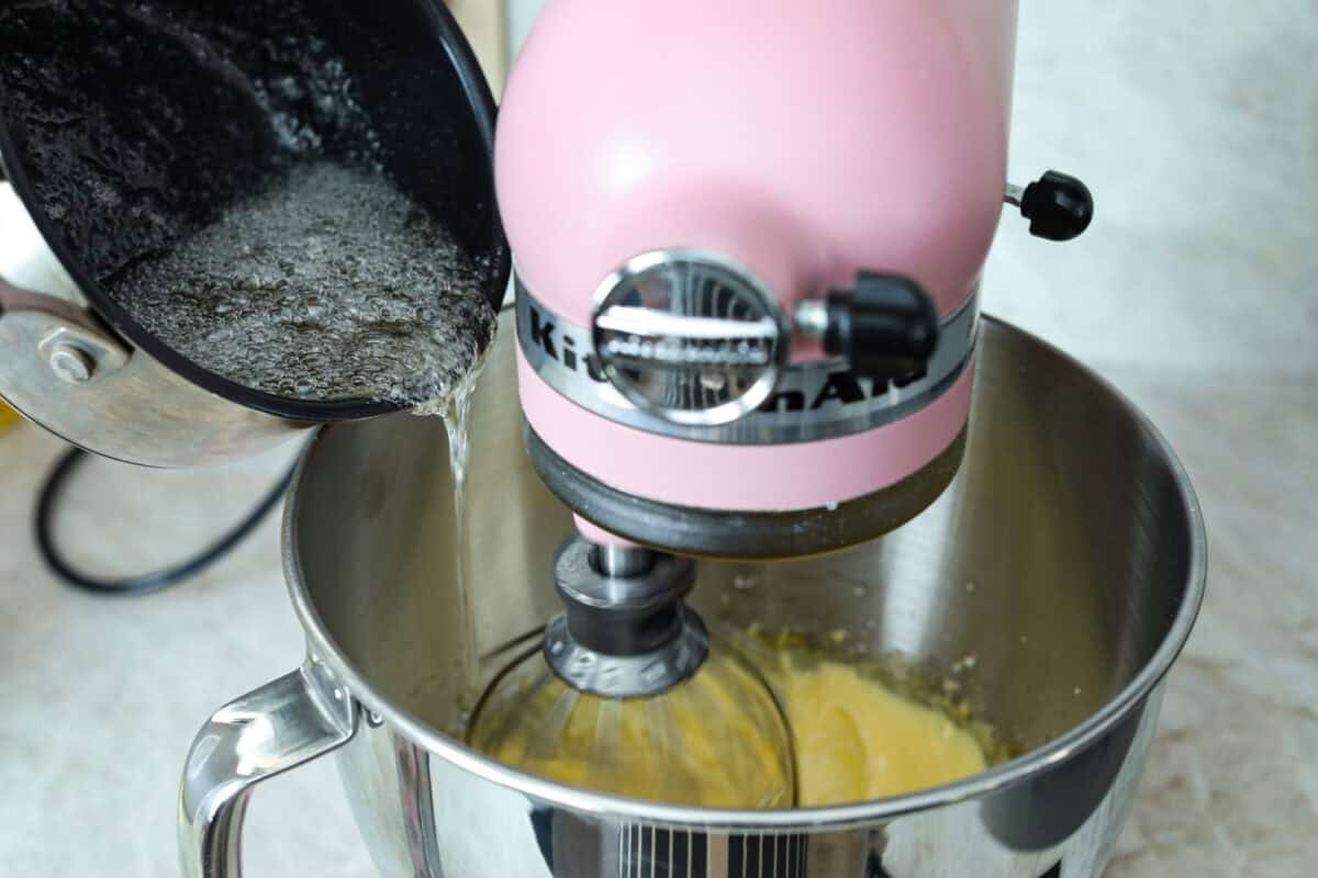 Angle shot of sugar syrup being slowly poured into a stand mixer with whipped egg whites. 