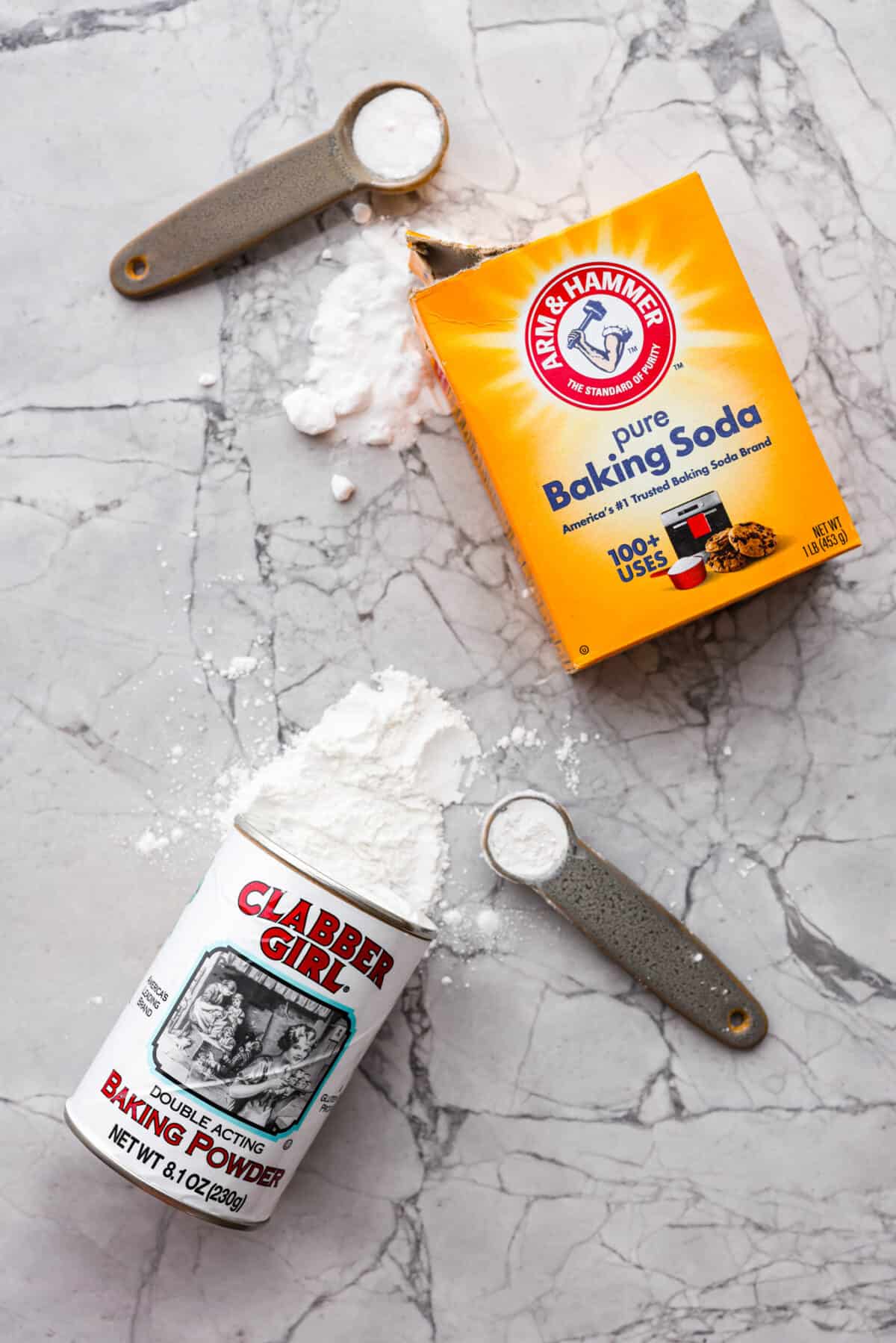 Top view of baking soda and baking powder spilling out of the containers with measuring spoons.