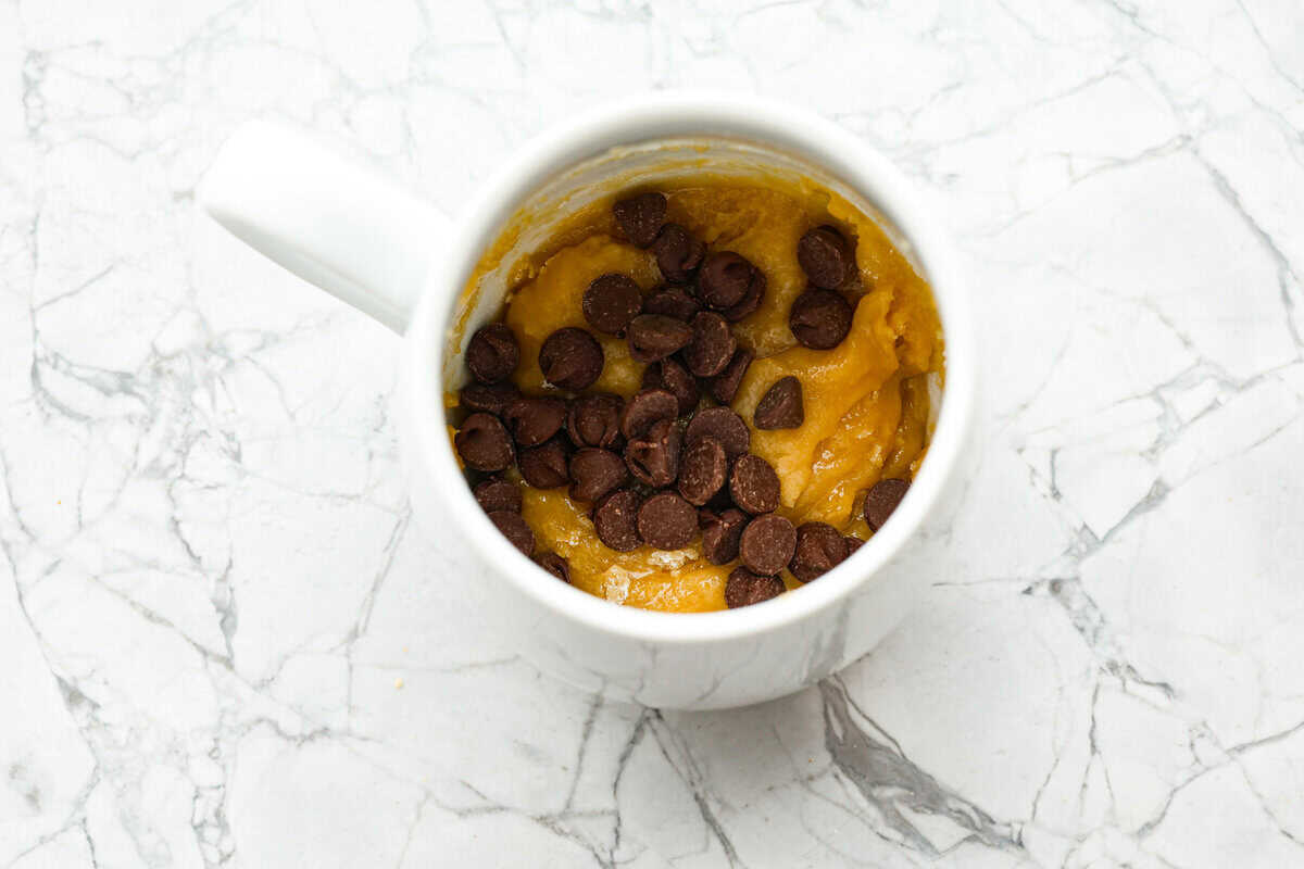 This cookie in a mug recipe is the perfect way to satisfy your cookie craving. In just a few minutes, you can enjoy a warm, buttery, ooey-gooey cookie right in your own mug. Top it with a scoop of ice cream or a dollop of whipped cream, and you'll find yourself in cookie heaven! Overhead shot of mug with cookie dough and chocolate chips dded.