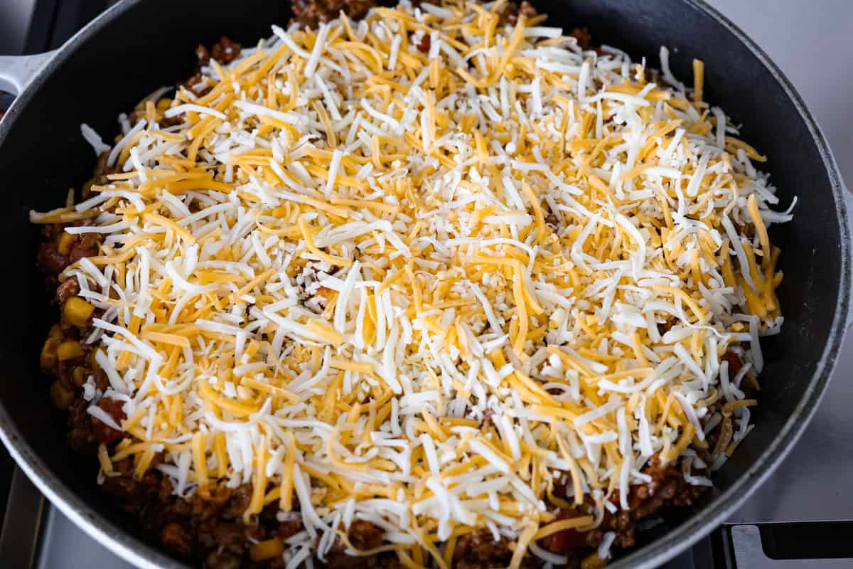 Cowboy Cornbread Casserole is a flavorful, comforting meal that's so easy to make. Seasoned ground beef, corn, pinto beans, and Rotel tomatoes are layered with shredded cheddar cheese and a cornbread topping for a winning dinner! Angle shot of ground beef, beans, and Rotel mixture with cheese sprinkled over the top of the inside of cowboy cornbread casserole .