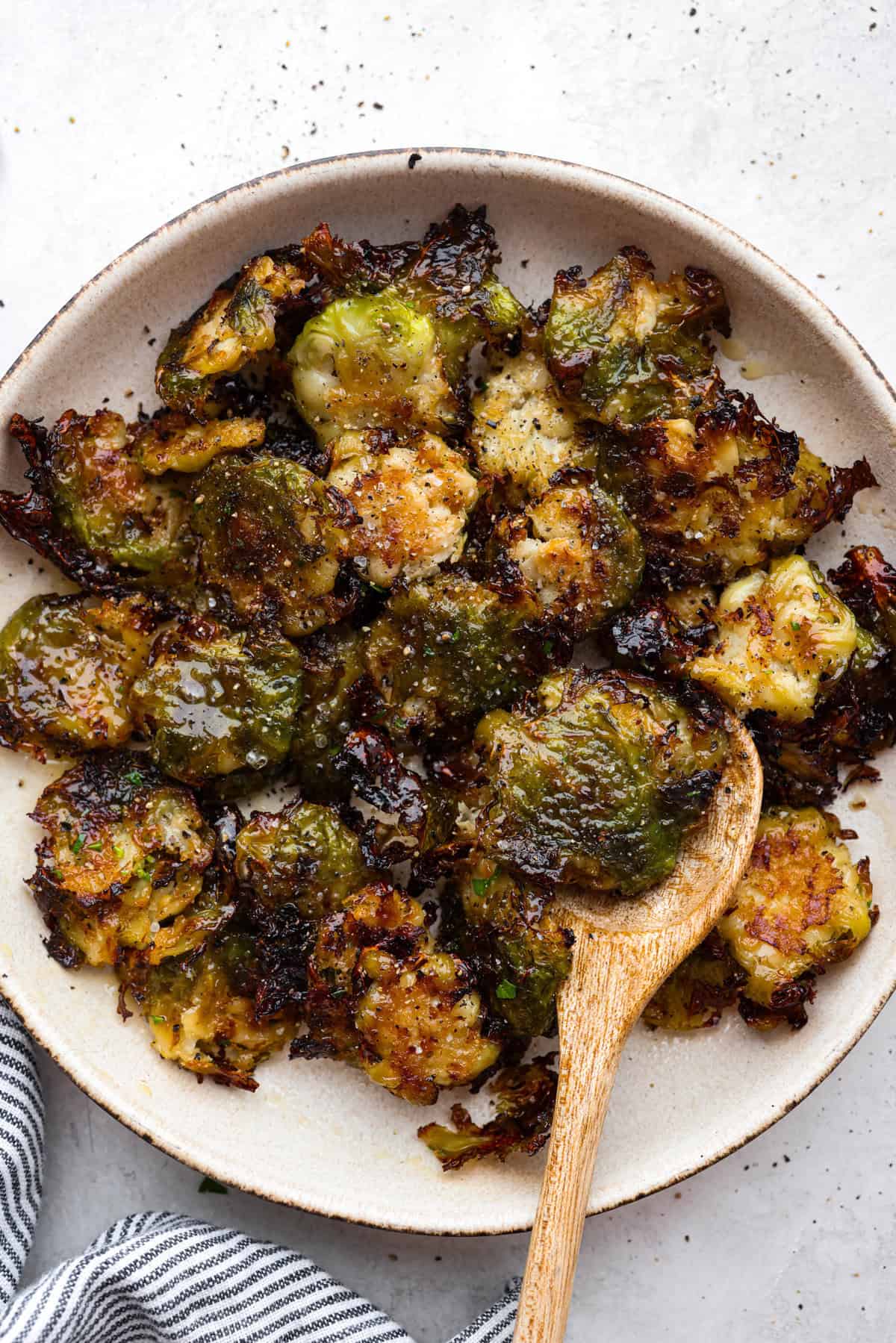 Irresistibly crispy Smashed Brussels Sprouts are simple to make, loaded with delicious flavor, and have the best crunchy texture. These pair well with almost any protein and will definitely upgrade your side dish game! Overhead shot of smashed Brussels sprouts on a plate with a wooden serving spoon.