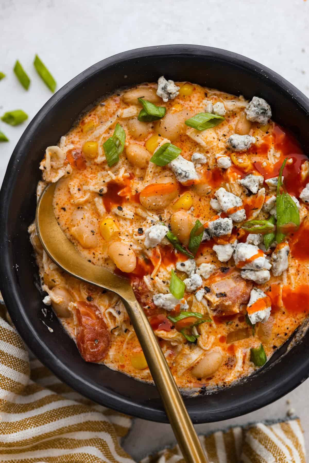 Top shot of a bowl of crockpot buffalo chicken chili with a golden spoon, topped with scallions, blue cheese, and a little hot suace. 