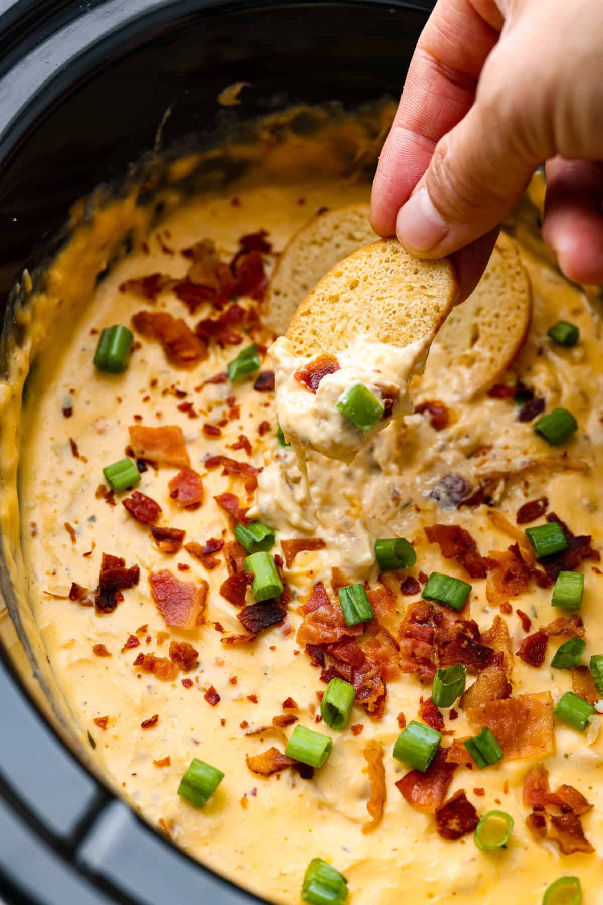 Close up shot of someone dipping a sliced baguette piece into the slow cooker crack chicken dip. 