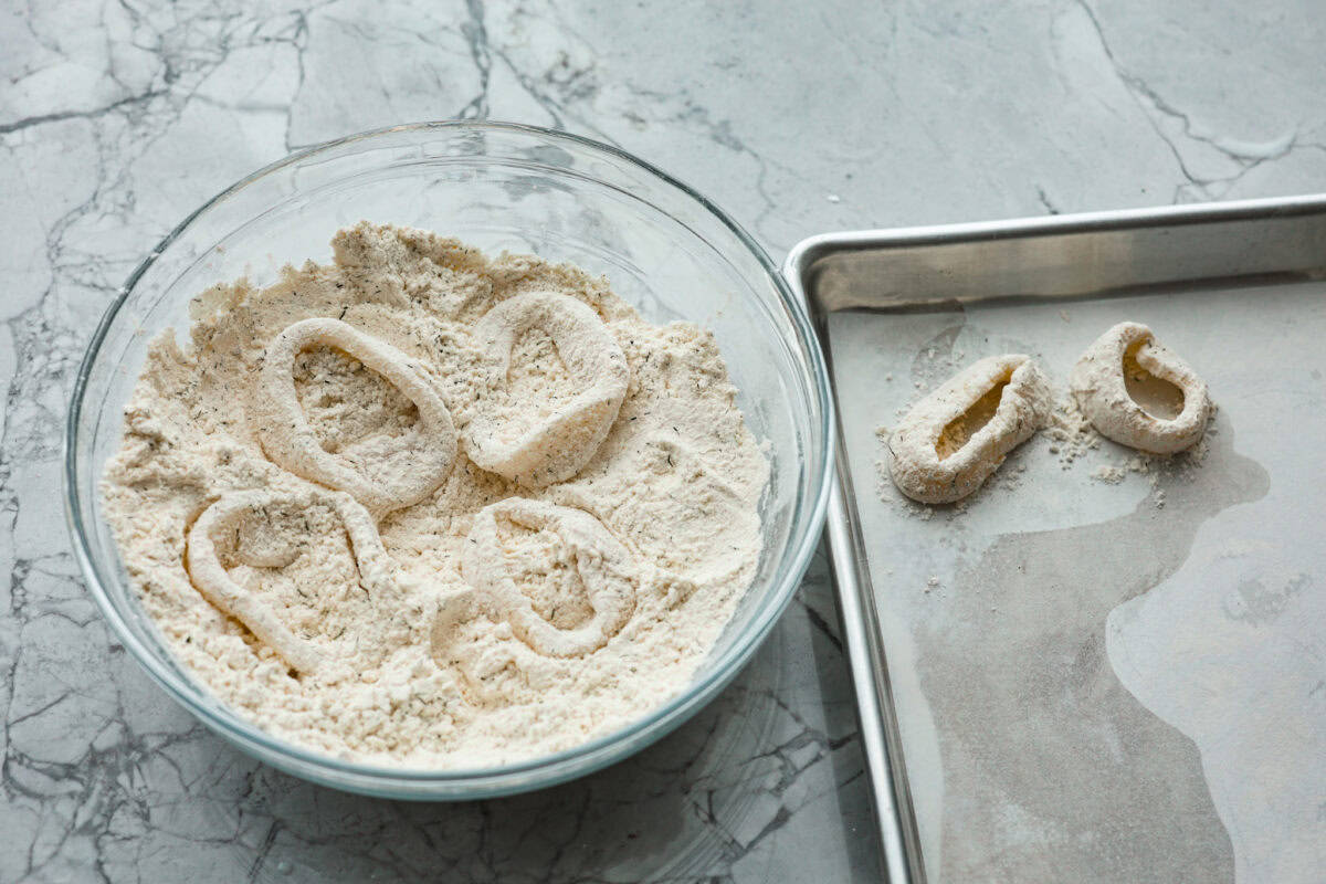 Overhead shot of calamari rings sitting in the flour and seasoning mixture in a bowl. With a couple of the dredged rings on a baking pan with parchment paper. 