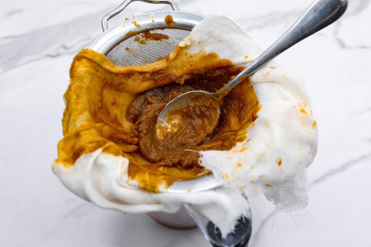 Overhead shot of a measuring cup with a wire strainer over the top covered in cheesecloth filled with puree mixture and a spoon. 