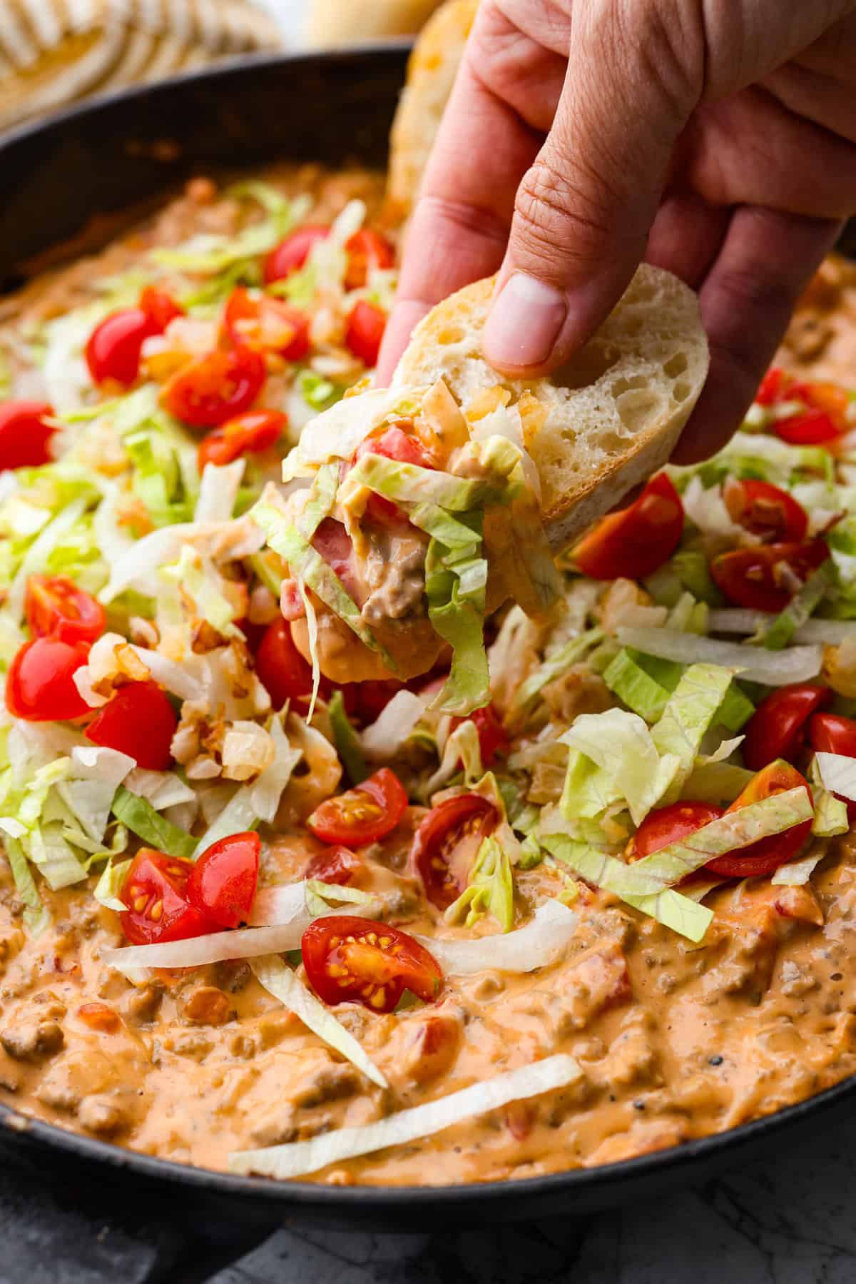 Cheeseburger dip is a crowd-pleasing appetizer for potlucks, parties, and get-togethers! This recipe takes delicious cheeseburger ingredients like beef, onion, tomato, relish, and cheese and turns them into a deliciously addicting snack! Close up shot of someone getting a bite of dip on a crust of bread.