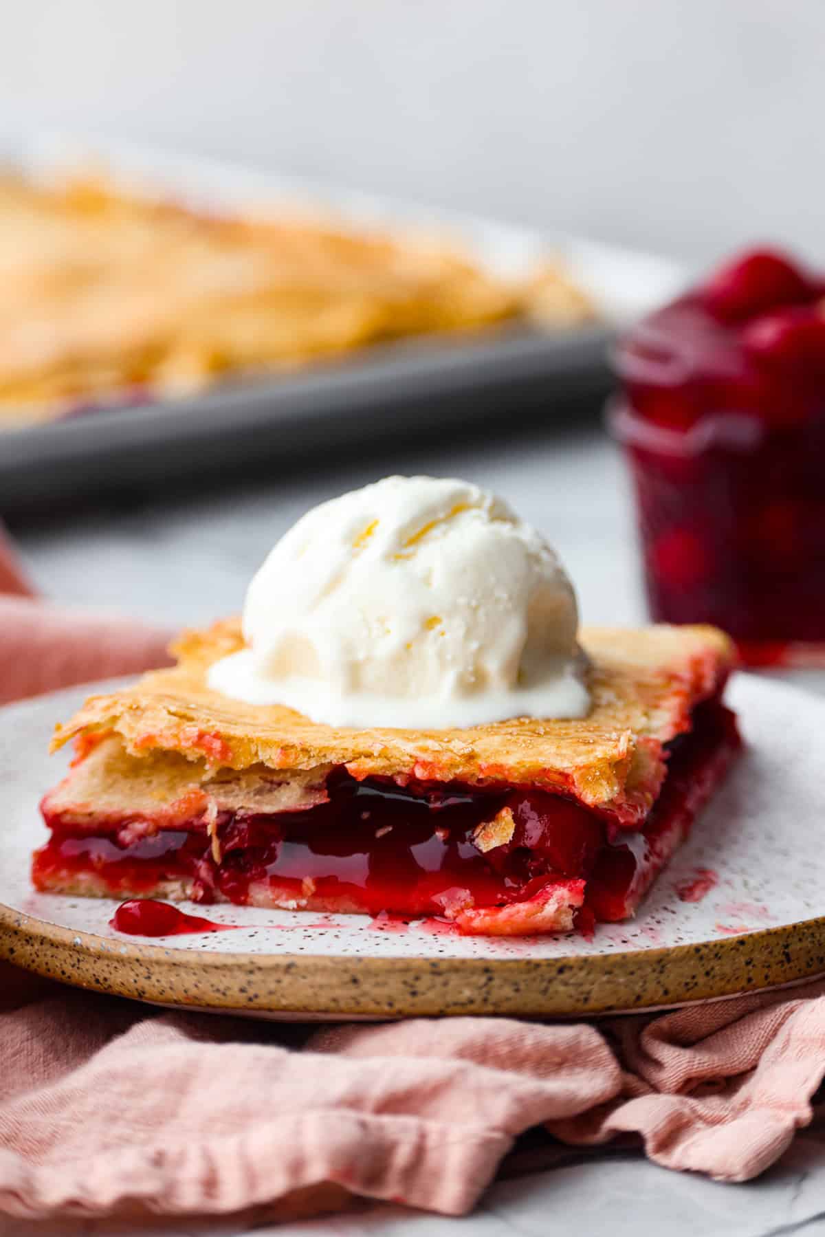 Cherry Slab Pie is an American classic made to serve a crowd! This recipe uses a tender, flaky crust that surrounds sweet, juicy cherry pie filling. It tastes incredibly delicious and is the perfect dessert for any time of year. Side shot of a plated slice of cherry pie slab with a scoop of vanilla ice cream on top.