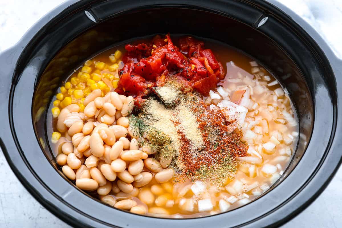 Crockpot buffalo chicken chili is a buffalo wing lover's dream. It has a tangy, fiery buffalo hot sauce base mixed with shredded chicken, smooth cream cheese, tender beans, and vegetables. If you love buffalo wings, you will love this chili! Overhead shot of the beans, corn, tomatoes, onion, buffalo sauce, chicken broth and seasonings in the crockpot.