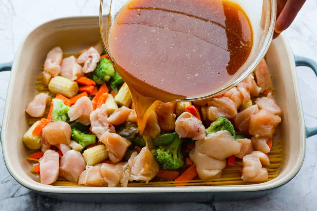 Overhead shot of someone pouring the sauce over the chicken, veggies and noodles in a baking dish.