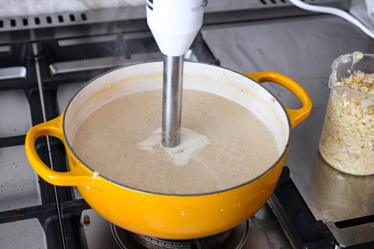 Angle shot of someone blending the soup with an immersion blender in the pot with a measuring cup with corn next to it. 