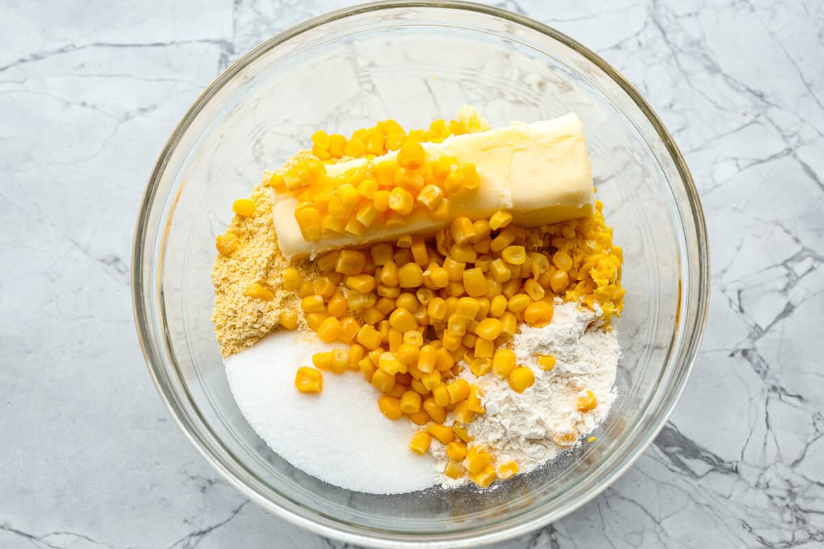Homemade Sweet Corn Tamale Cakes are an explosion of delicious flavor in every bite! With this easy recipe you can bring this Cheesecake Factory favorite right to your own kitchen. Add the signature topping to really take these to the next level! Overhead shot of pulverized corn, whole corn, flours, sugar, butter, and salt in a glass bowl.