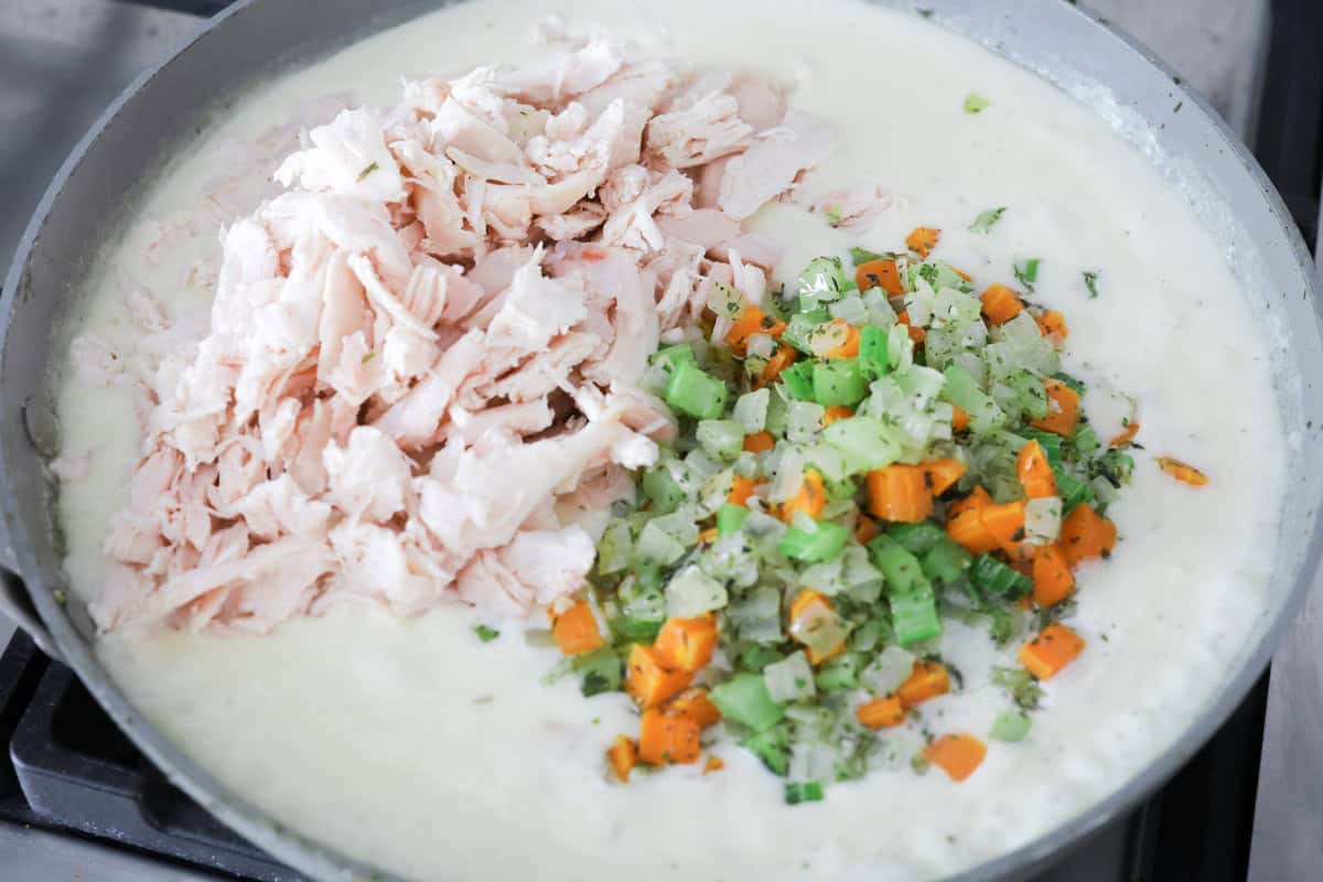 Chicken pot pie casserole is a fun twist on the classic pot pie. It's made with buttery biscuit dough, tender chicken, crunchy veggies, and the most delicious creamy sauce loaded with incredible seasoning. Comfort food at its finest! Angle shot of the veggies and chicken being added to the cream sauce in a skillet.