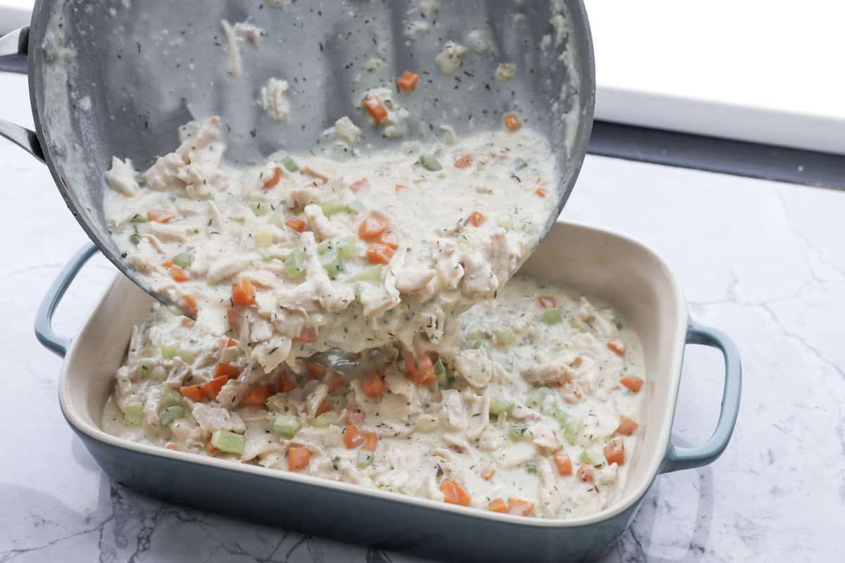 Chicken pot pie casserole is a fun twist on the classic pot pie. It's made with buttery biscuit dough, tender chicken, crunchy veggies, and the most delicious creamy sauce loaded with incredible seasoning. Comfort food at its finest! Angle shot of someone pouring the veggie, chicken and sauce mixture into a baking dish.