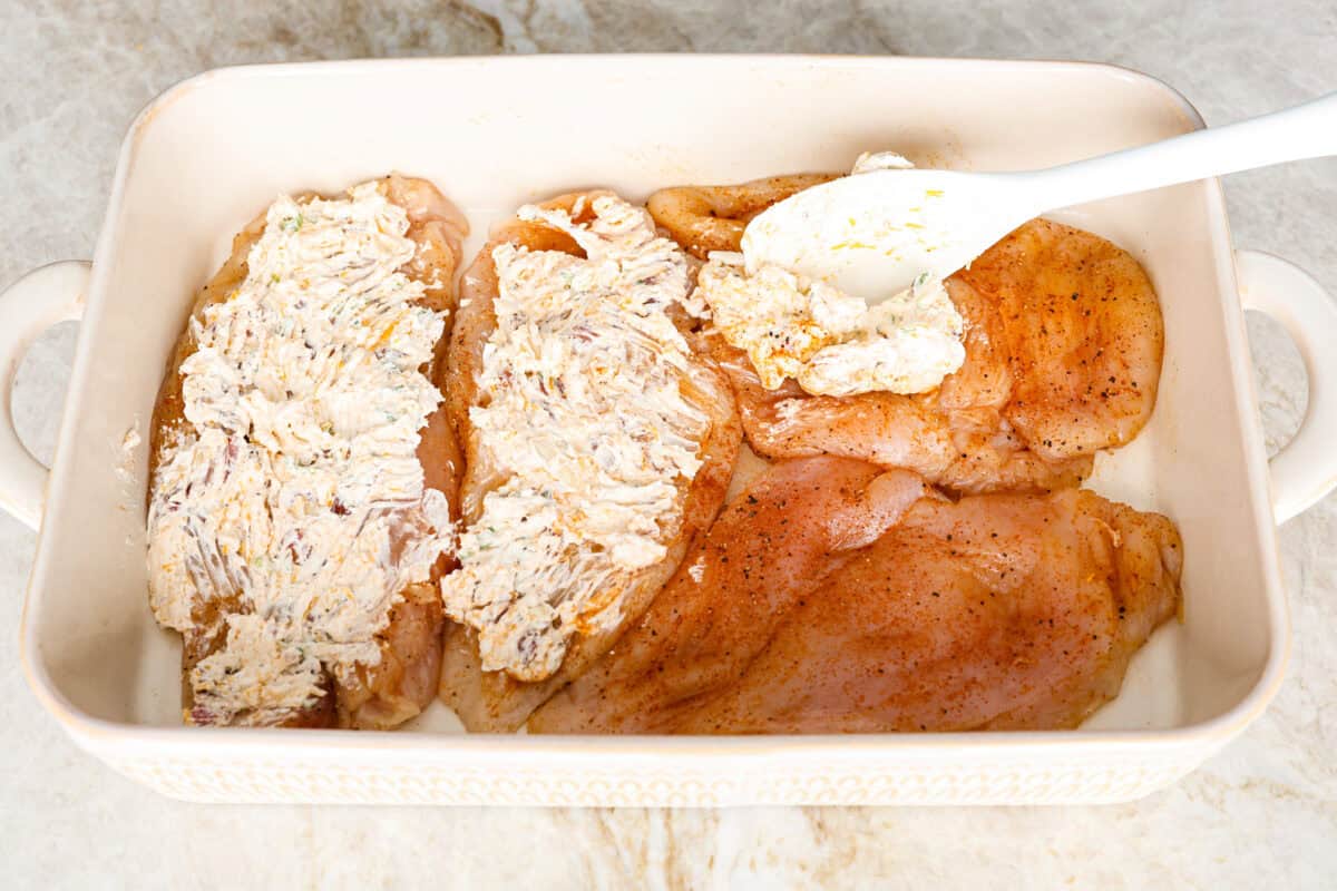 Angle shot of someone covering the seasoned chicken breasts with the cream cheese topping mixture in a baking dish. 