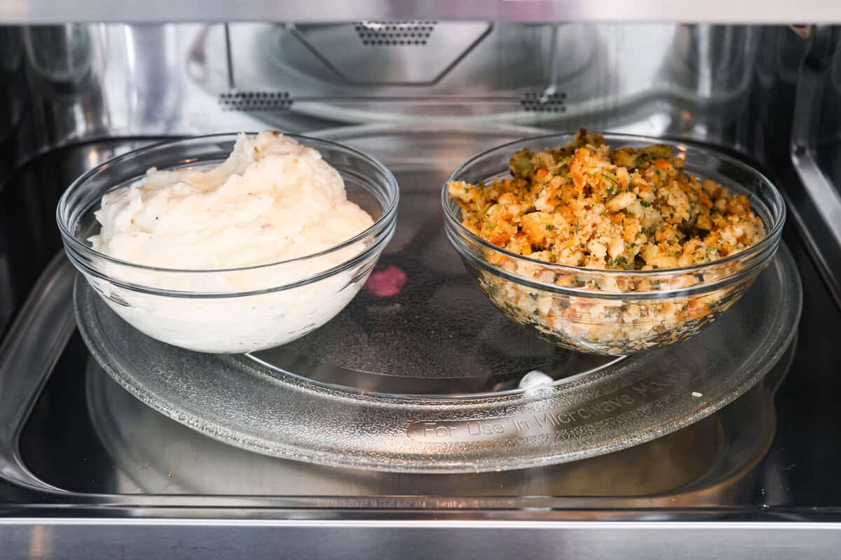 Turkey Stuffing Balls capture the taste of Thanksgiving in a single bite! Made with tender turkey, flavorful stuffing, and mashed potatoes. They’re rolled into perfectly poppable bites and served with rich gravy for dipping. Side shot of a bowl of mashed potatoes next to a bowl of stuffing in a microwave.